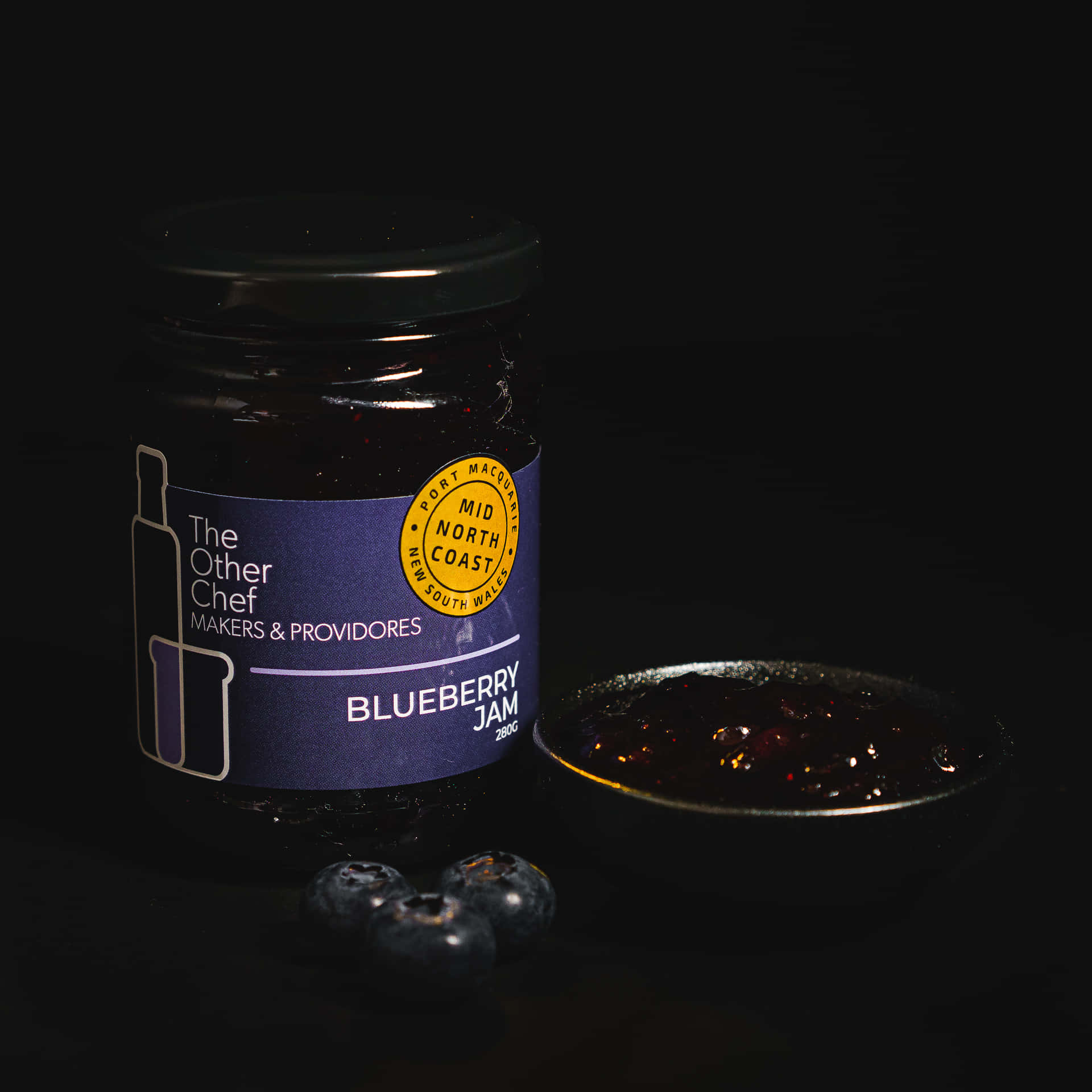 Blueberry Jam 桌布 壁紙 — 一樽藍莓果醬，旁邊有新鮮莓果和一匙果醬，在黑色背景下戲劇性地打光。
