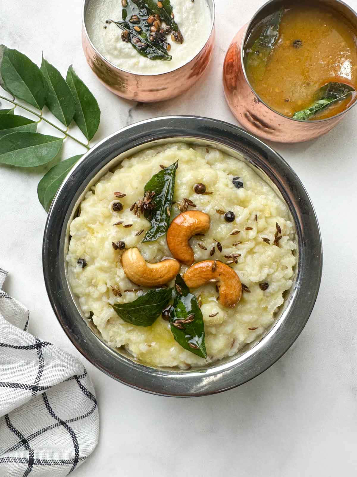 Pongal 桌布 壁紙 — 一款奶油米飯，上面放有腰果、咖喱葉和孜然籽，盛載於金屬碗中，搭配銅杯和綠葉，置於白色表面上。