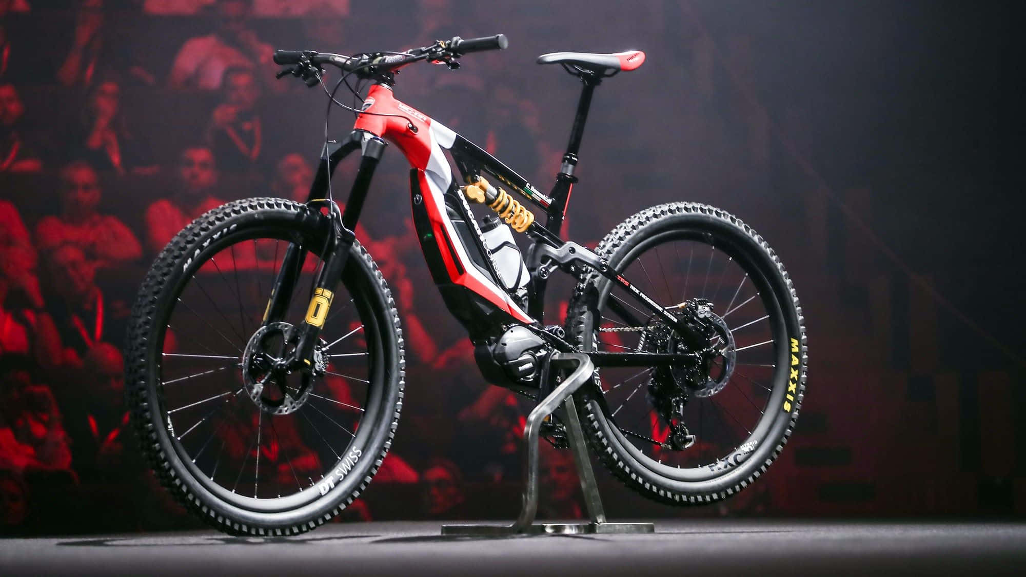 Electric Bike 桌布 壁紙 — 一款時尚的紅黑電動山地車在產品發佈會上展示，背後有紅色燈光下的模糊觀眾。