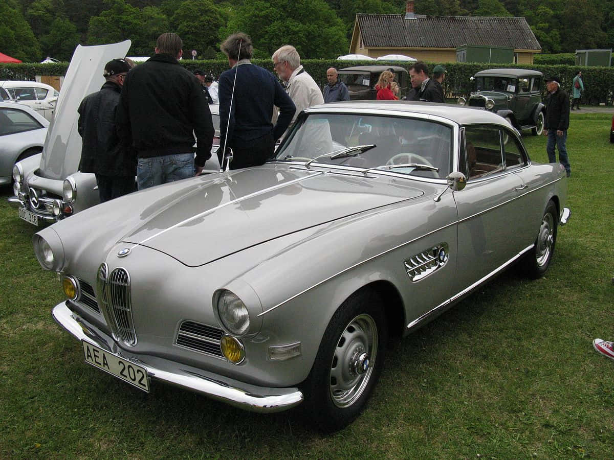 一款流線時尚的BMW 壁紙 — 一架流線型銀色復古BMW 507跑車在戶外經典車展的日光下閃耀，周圍有觀眾聚集。