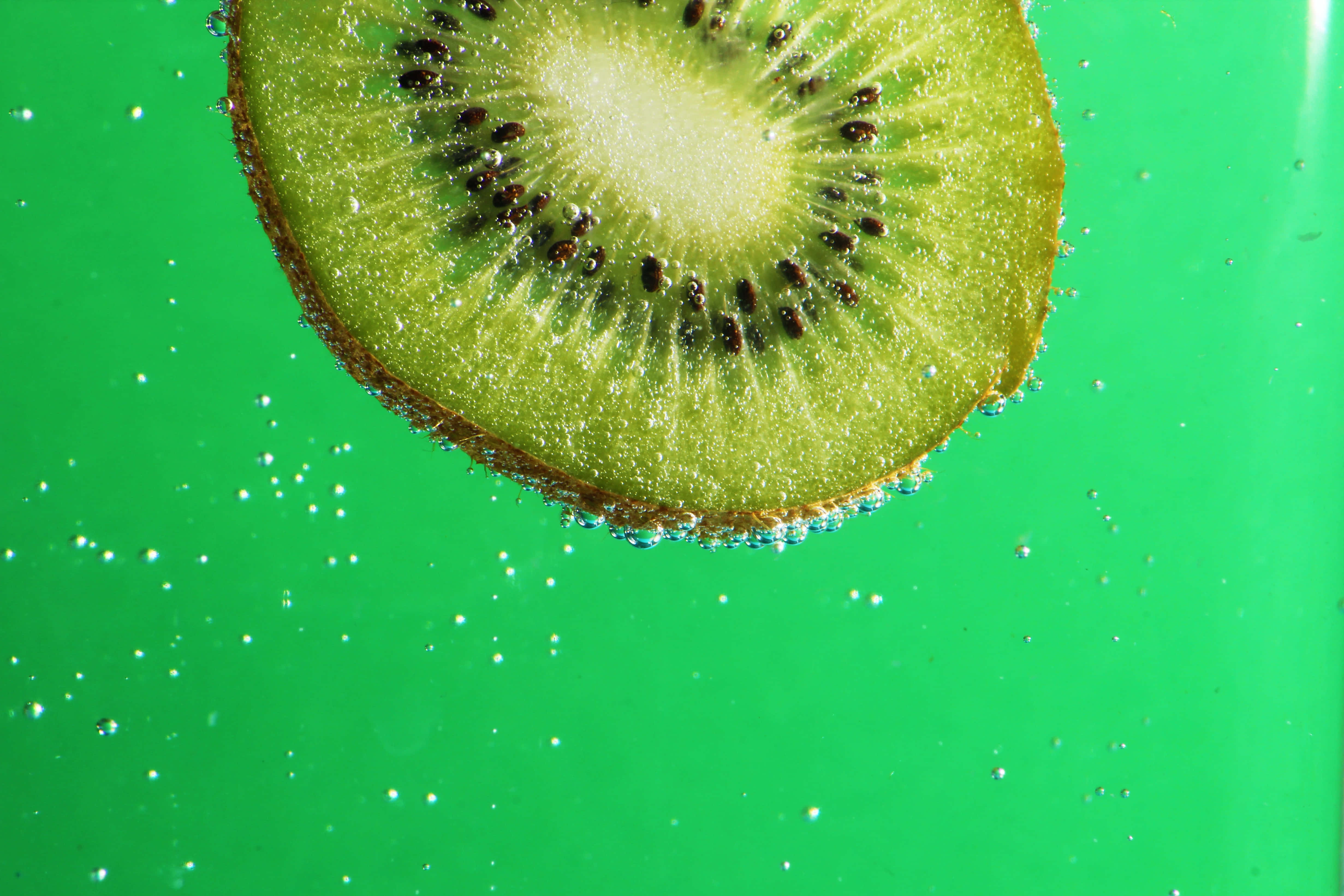 Kiwi 桌布 壁紙 — 一片充滿活力的綠色奇異果片漂浮在水中，周圍環繞著閃閃發光的氣泡，背景是鮮豔的綠色。