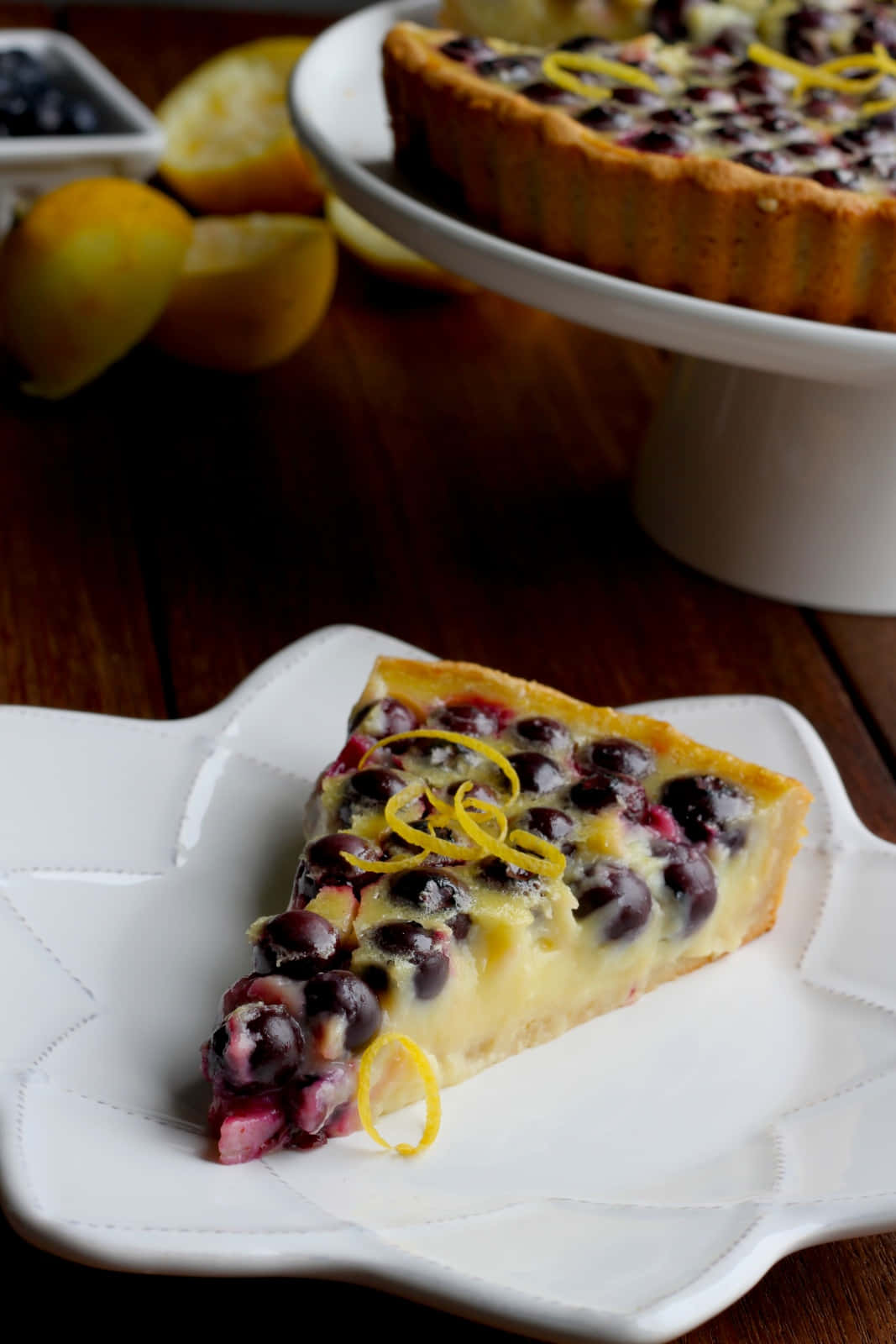 Blueberries Tart 桌布 壁紙 — 一片藍莓檸檬撻，飾以檸檬皮，放在白色碟上，旁邊有整個檸檬和一個放在架上的完整撻。