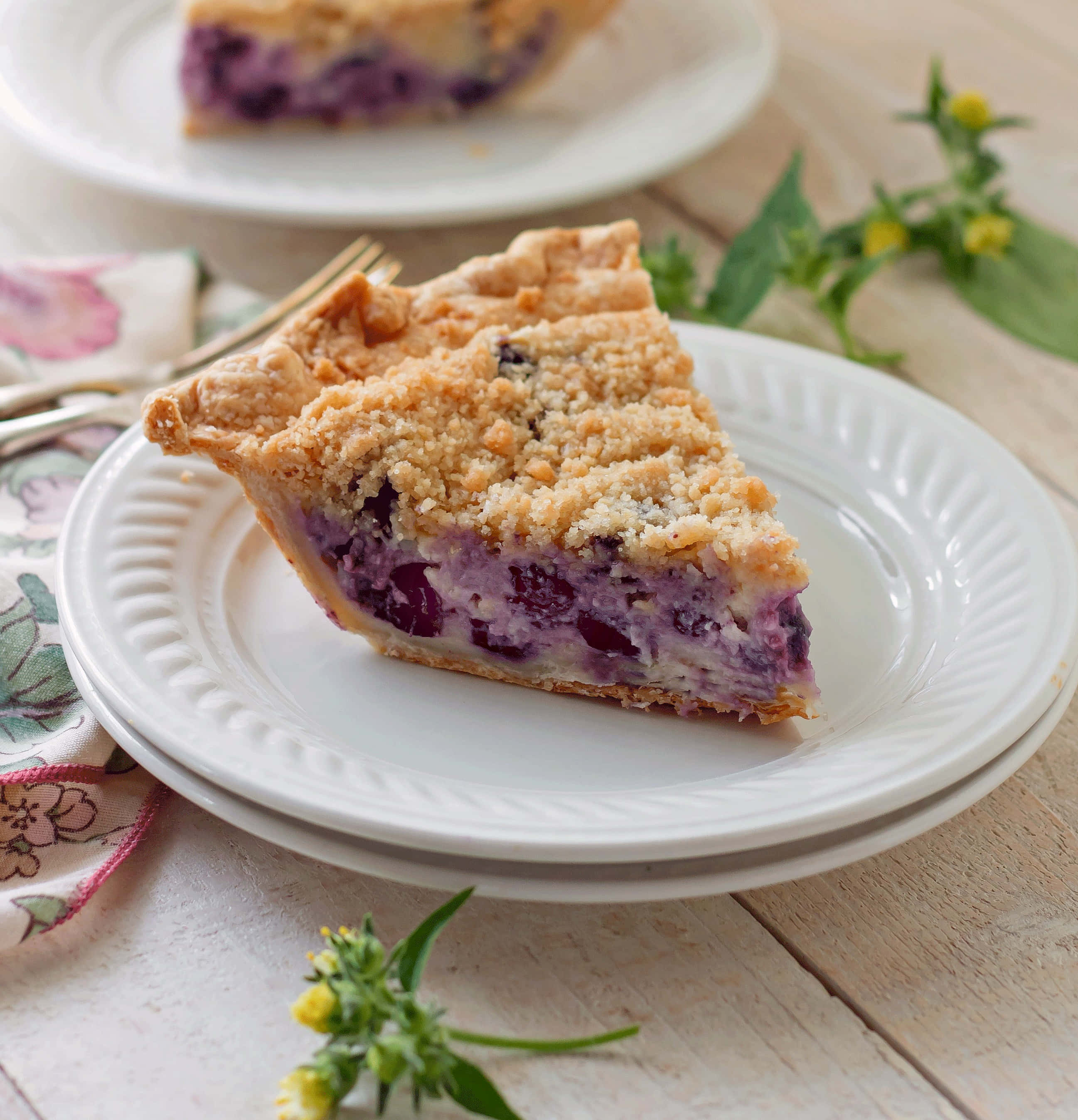 Blueberry Pie 桌布 壁紙 — 一片金黃脆皮的藍莓金寶批放在白色碟子上，置於鄉村風木桌上，旁邊有新鮮香草和花卉圖案餐巾。