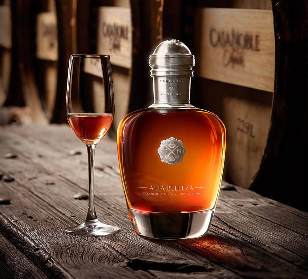 Casa Noble Alta Belleza 壁紙 — 一瓶 Alta Belleza 龍舌蘭酒和一個玻璃杯放在質樸的木桌上，旁邊是威士忌酒桶。