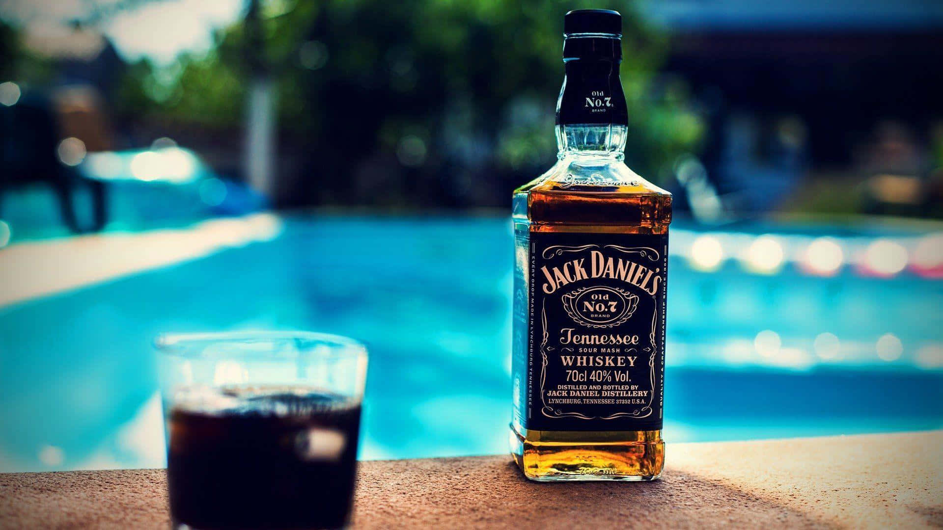 alcohol 桌布 壁紙 — 一瓶Jack Daniel's田納西威士忌和一杯加冰的威士忌放在泳池邊的 ledge 上，背景是模糊的藍色泳池。