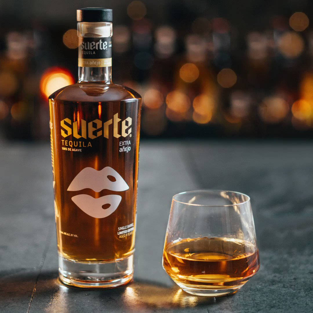 Suerte Anejo「Lucky Lips」 壁紙 — 一瓶Suerte Extra Añejo龍舌蘭酒和一杯琥珀色液體放在深色表面上，背景是溫暖模糊的酒吧。