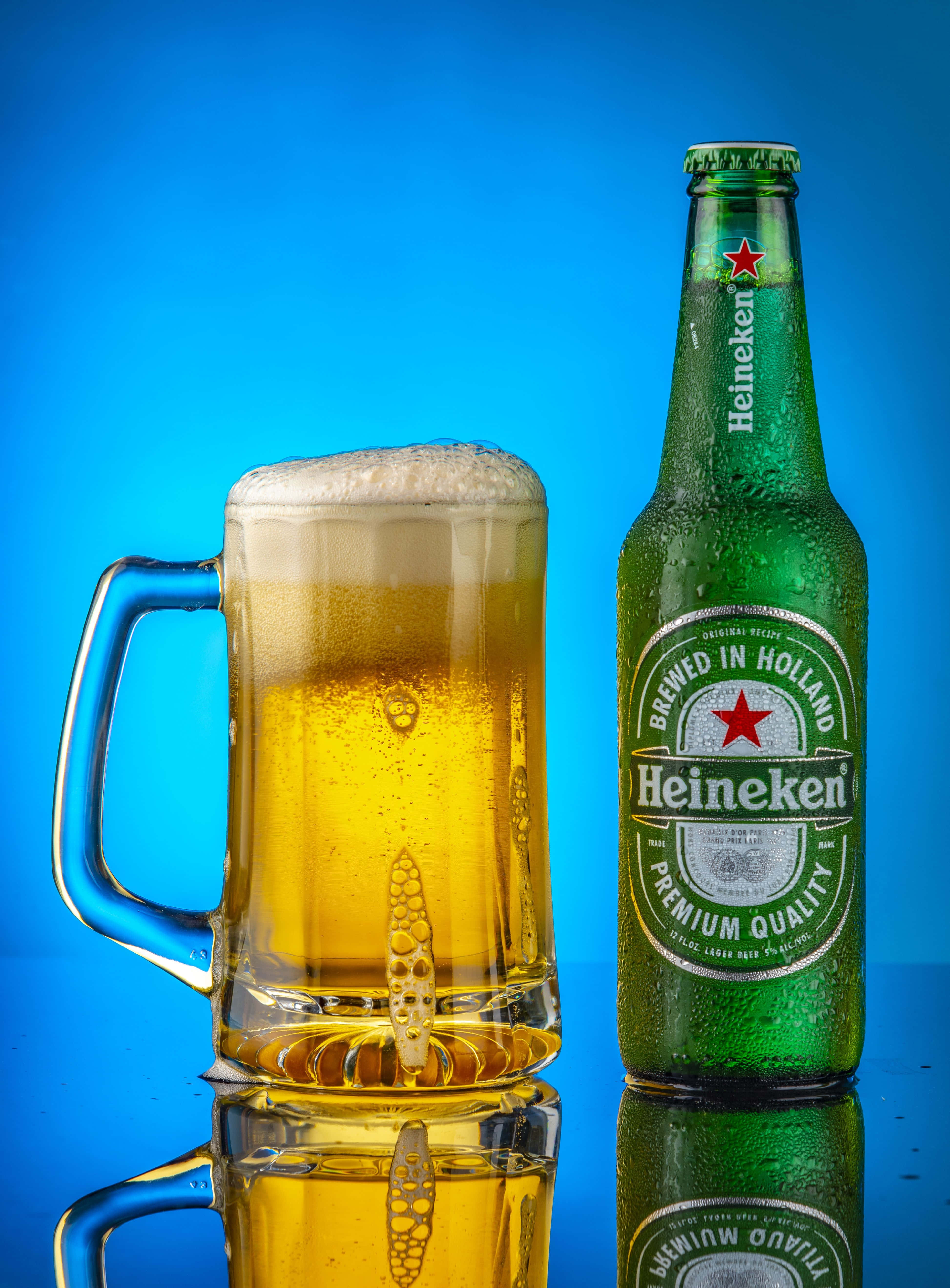 Heineken 桌布 壁紙 — 一瓶冰凍的喜力啤酒和起泡的啤酒杯在光亮的藍色表面上反射，背景是鮮豔的藍色。