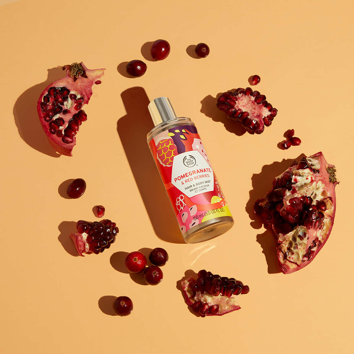 Red Fruit 桌布 壁紙 — 一瓶石榴和紅莓頭髮及身體噴霧，周圍散落著石榴籽和完整莓果，放在溫暖的米色背景上。