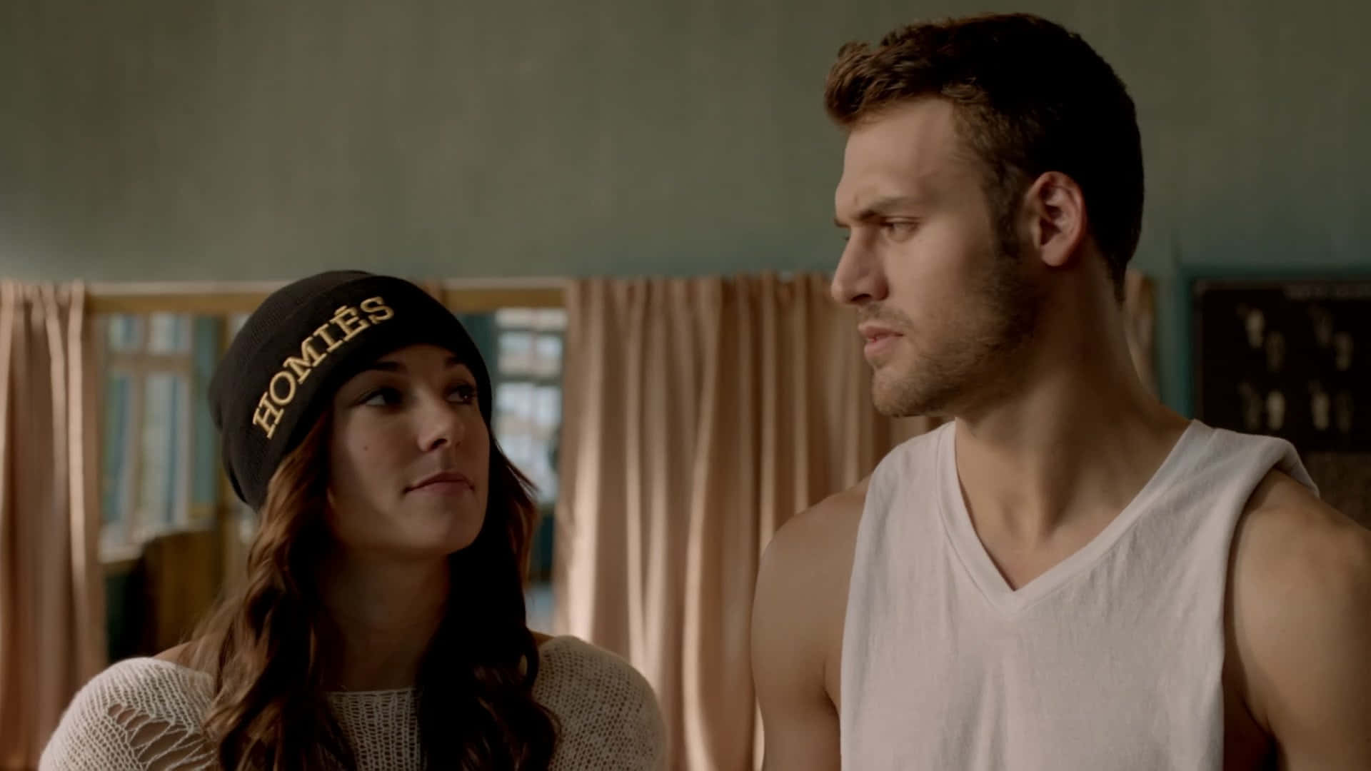 Briana Evigan 桌布 壁紙 — 一男一女站在燈光柔和的房間裡，女子戴著「HOMIES」冷帽，彼此靜靜地凝視對方。