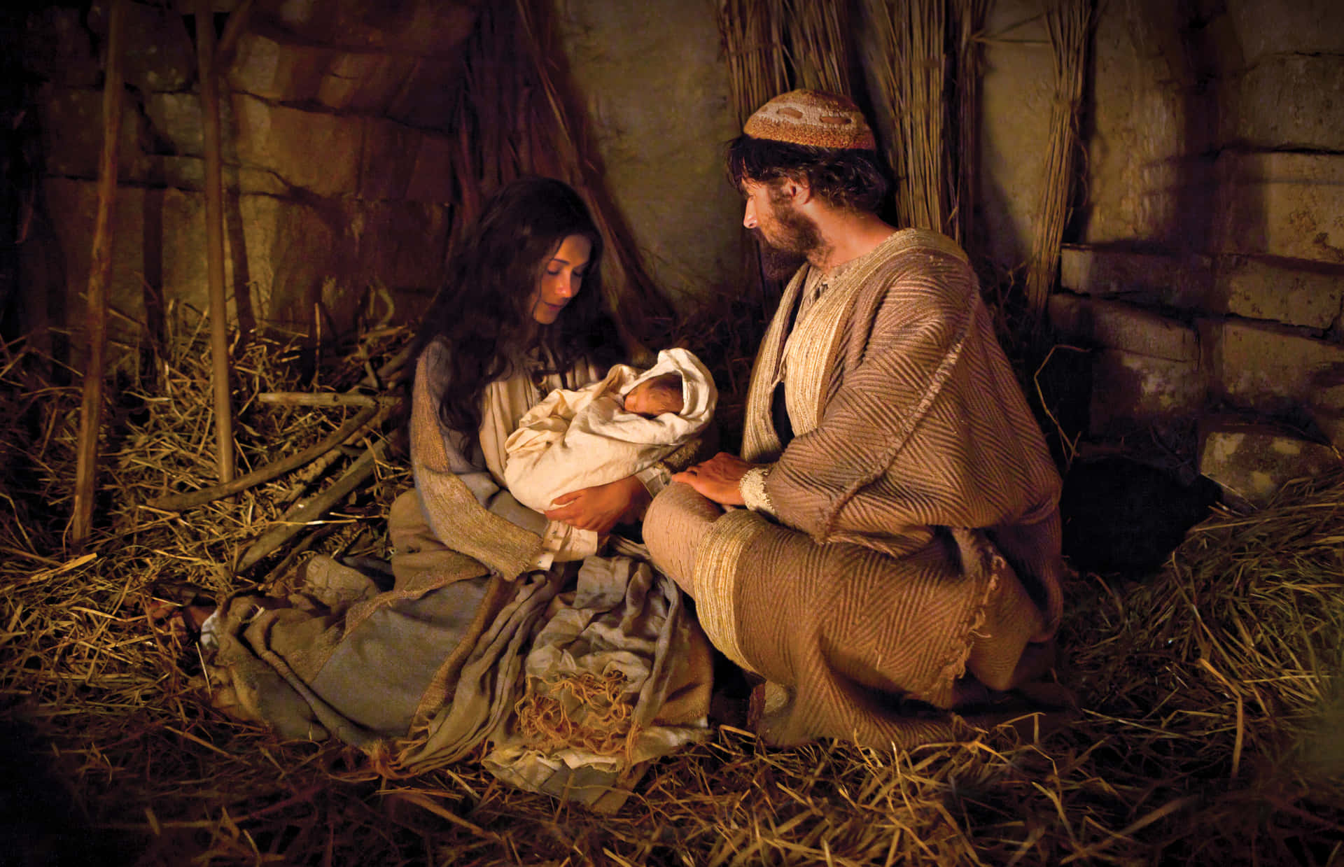 Nativity Scene 桌布 壁紙 — 一男一女身穿古裝長袍，謙卑地跪在馬槽裡，懷中抱著一個用襁褓包裹的新生嬰兒，周圍是稻草和石牆。