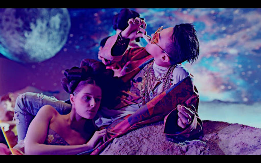 G-DRAGON《Bae Bae》 壁紙 — 一男一女躺臥在岩石上，下方是宇宙天空，有巨大的月亮和星雲，閃爍著鮮豔的紫色和藍色。