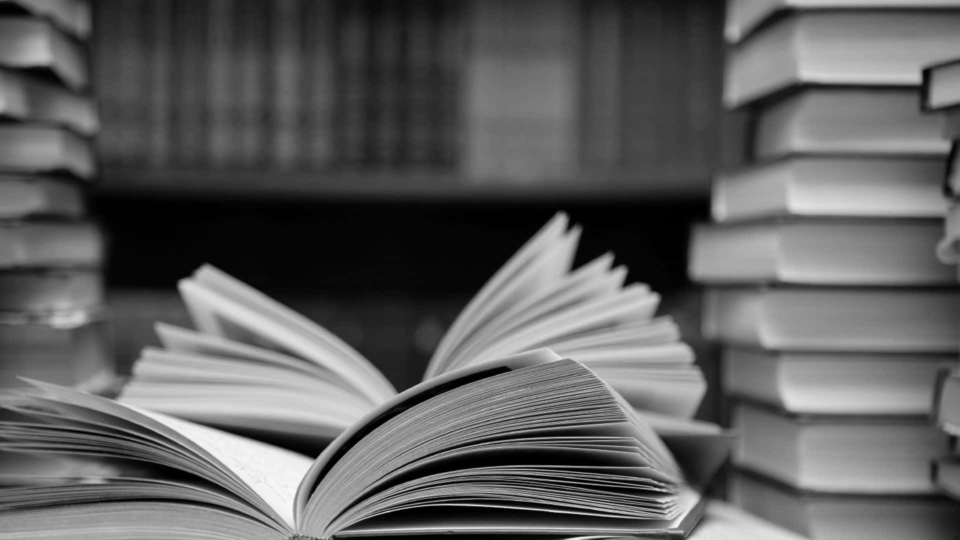 一疊書籍的單色藝術照，投下戲劇性陰影 壁紙 — 一本單色打開的書本放在寧靜圖書館書架上高聳的書堆之間。