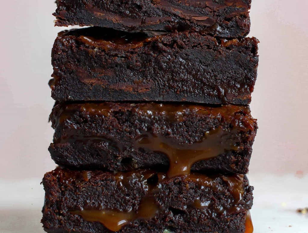 Caramel Brownie 桌布 壁紙 — 一疊濃郁軟心的布朗尼蛋糕，黏稠的焦糖在層與層之間滲出，襯以柔和粉紅背景。