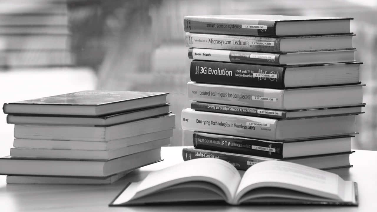 Black And White Book 桌布 壁紙 — 一疊關於無線和微系統技術的技術書籍整齊地放在桌子上，前景有一本打開的書。