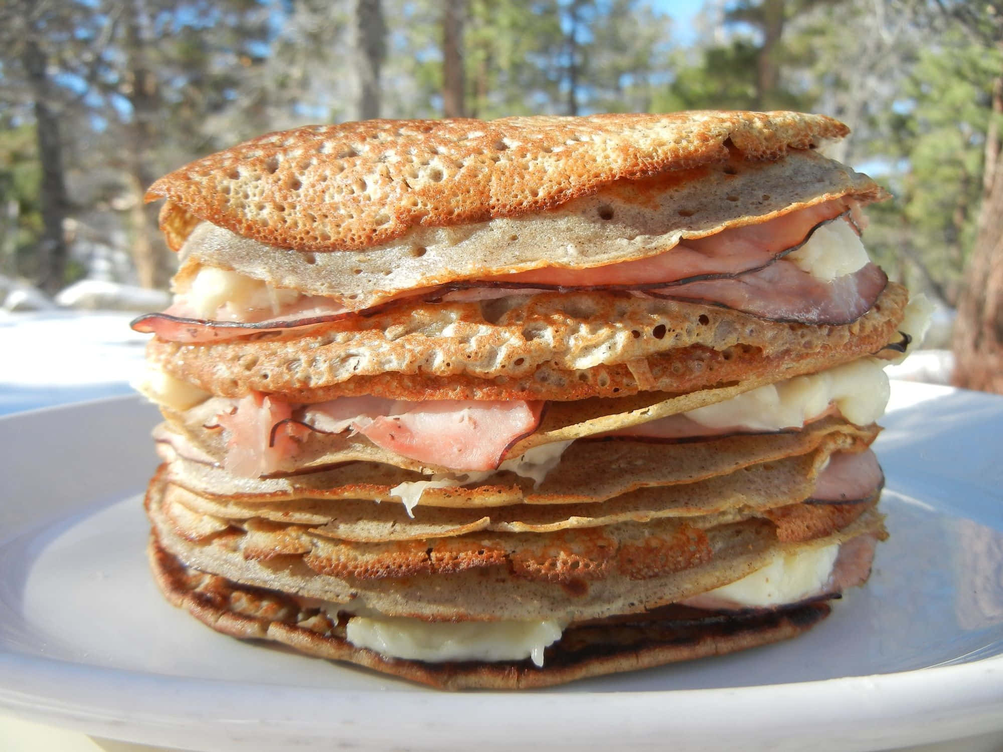 Black Forest Ham 桌布 壁紙 — 一疊高高的金黃色可麗餅，內餡是融化的芝士和火腿，放在白色碟子上，在戶外明亮的陽光下。