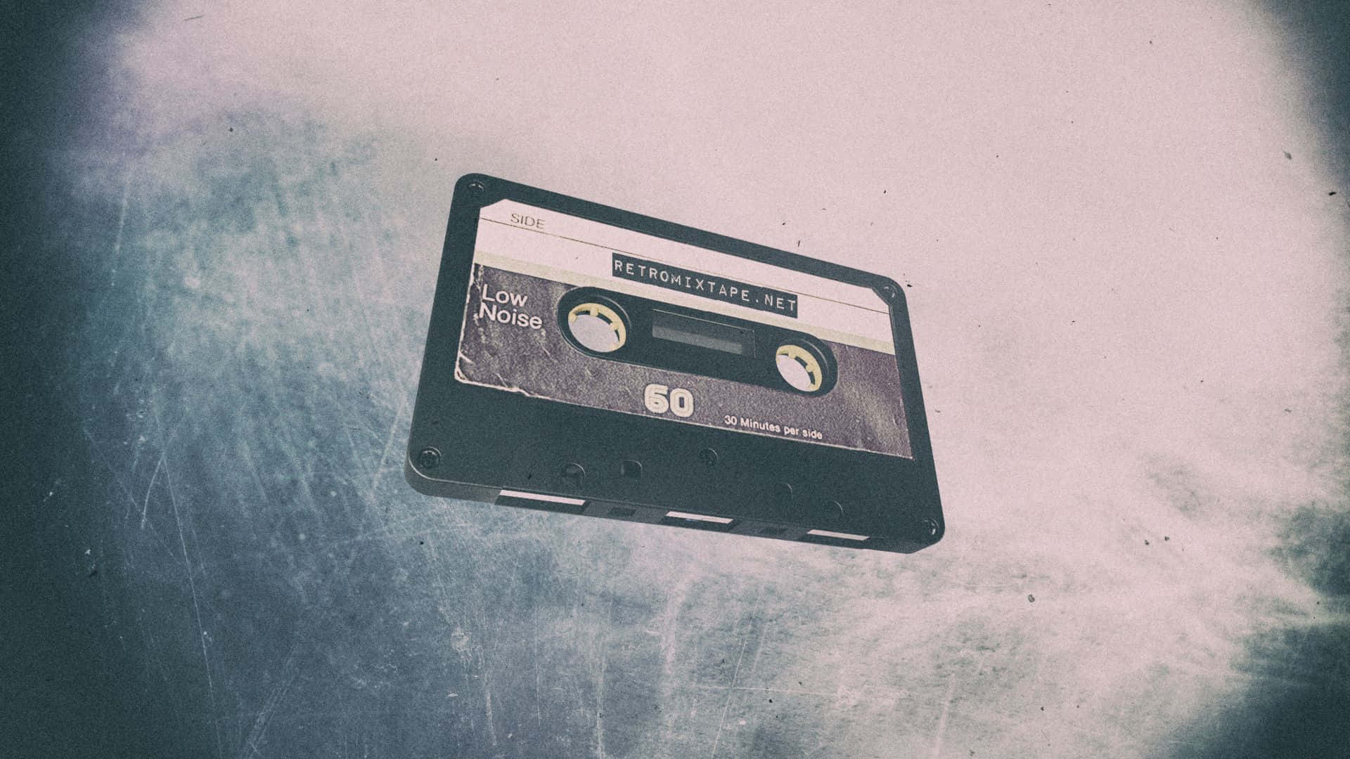 一盒盒磁帶，寫下音樂歷史 壁紙 — 一個標有「RETROMIXTAPE.NET」的復古黑色卡式錄音帶，放在一個有刮痕的金屬表面上，背景是褪色的藍白漸變色。