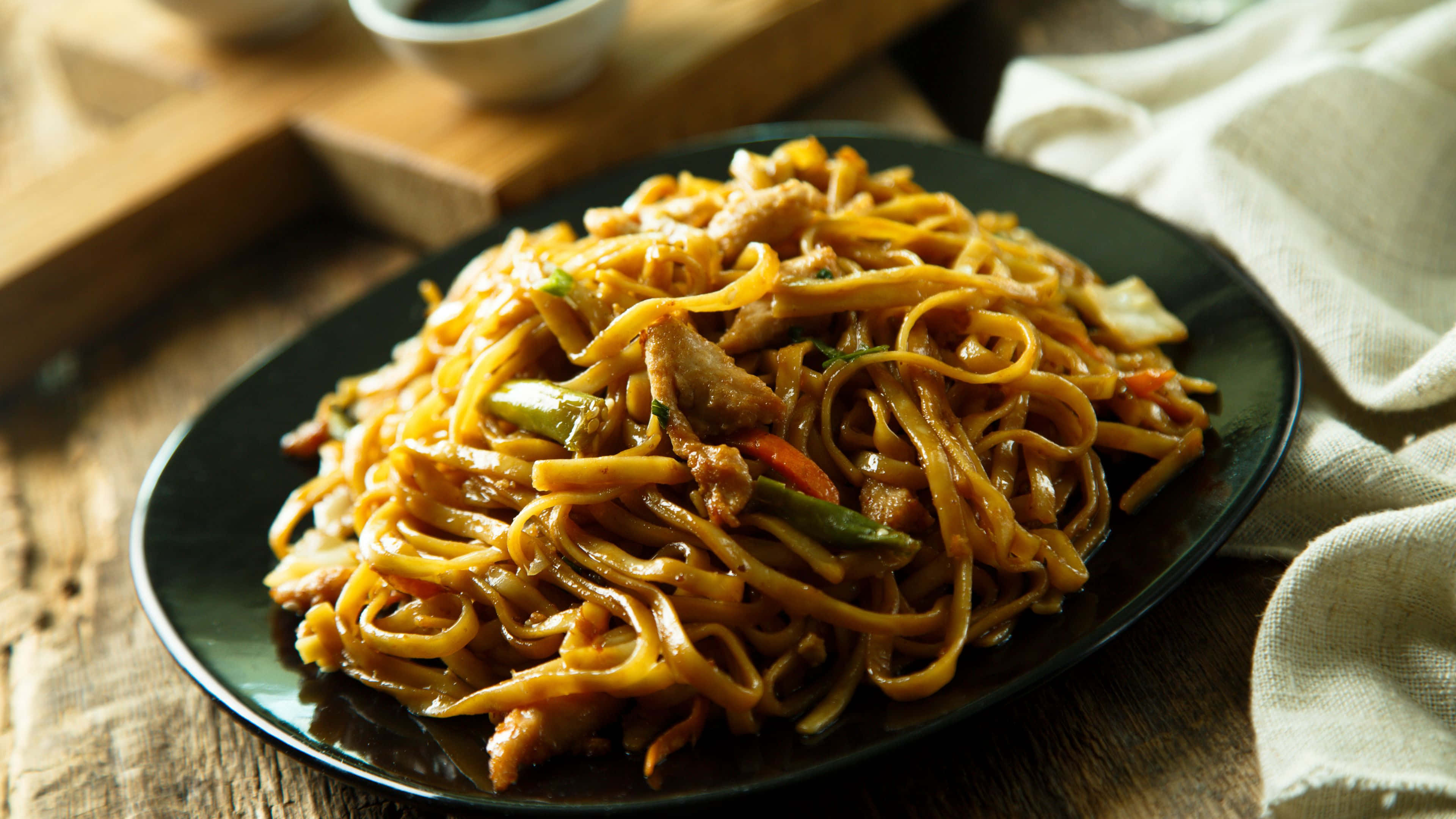 Noodles 桌布 壁紙 — 一盤冒著熱氣的炒麵，配有肉類和蔬菜，放在質樸的木桌上。
