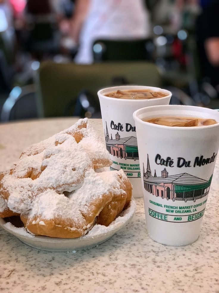 Blue Bayou 桌布 壁紙 — 一盤撒滿糖粉的貝涅餅旁邊有兩杯 Café Du Monde 的凍咖啡，放在有斑點的枱面上。