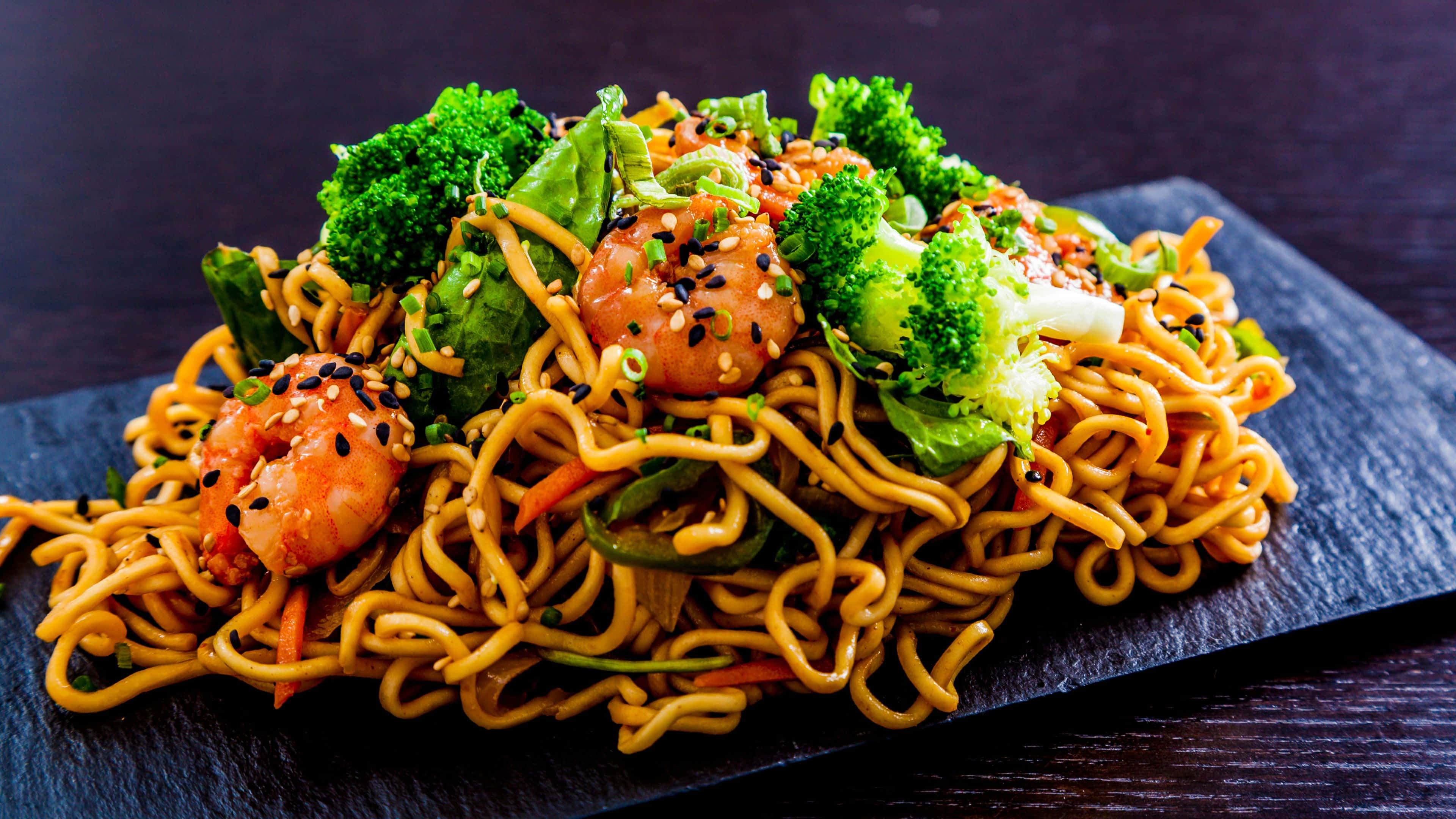Noodles 桌布 壁紙 — 一盤色彩繽紛的炒金黃麵條，配上蝦仁、西蘭花和芝麻，高高堆在深色石板盤上。