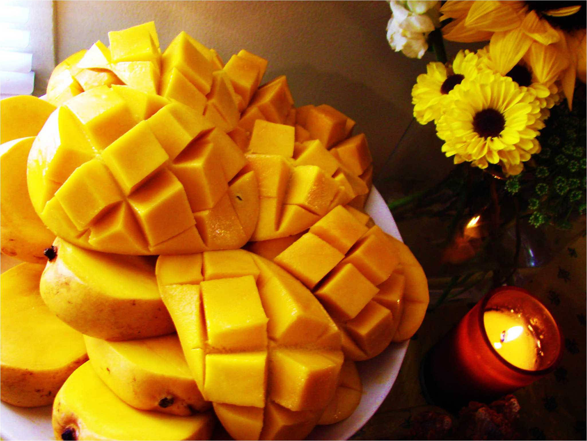 Mango 桌布 壁紙 — 一盤鮮豔的芒果粒放在向日葵花瓶和發光蠟燭旁邊，在溫暖的環境光中。
