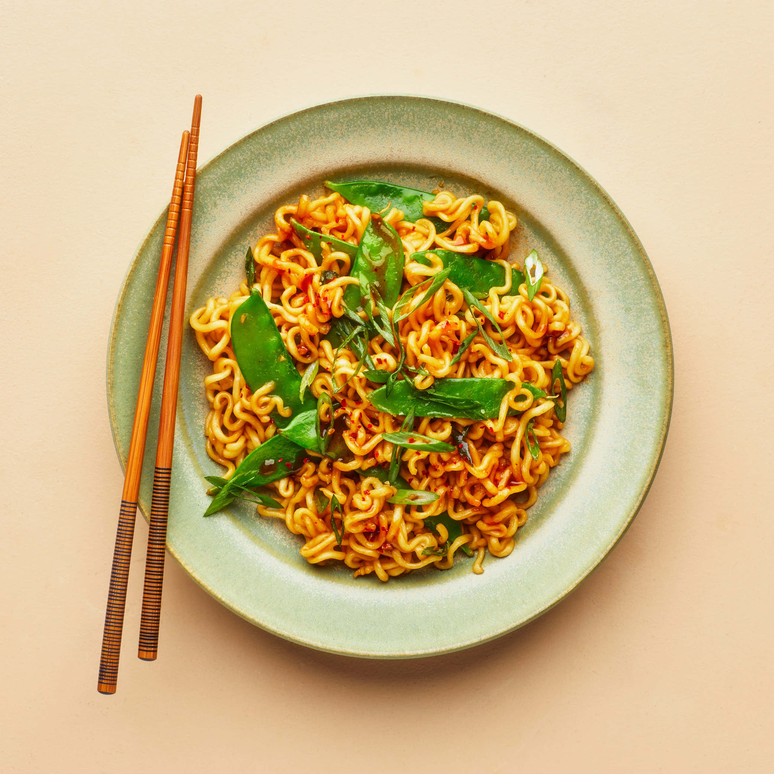 Noodles 桌布 壁紙 — 一碗充滿活力的橙色拉麵，上面放上新鮮的綠色荷蘭豆和紅辣椒油，配上木筷子，放在柔和的綠色碟子上。