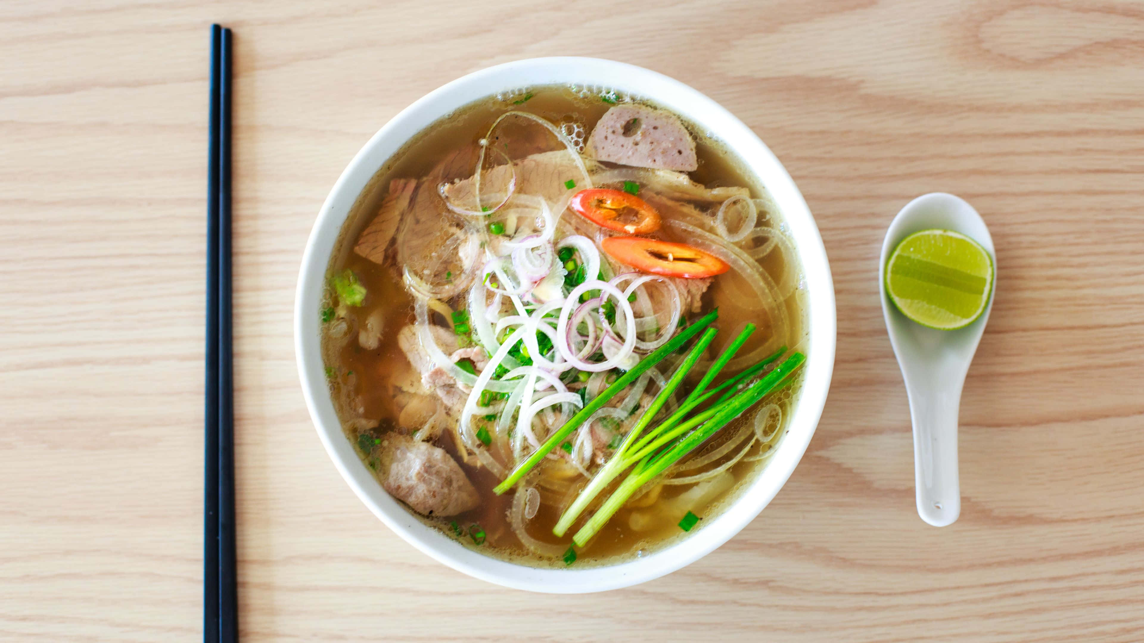 Noodles 桌布 壁紙 — 一碗冒煙的越南河粉，配有米粉、肉片、青檸和香草，放在淺色木桌上。