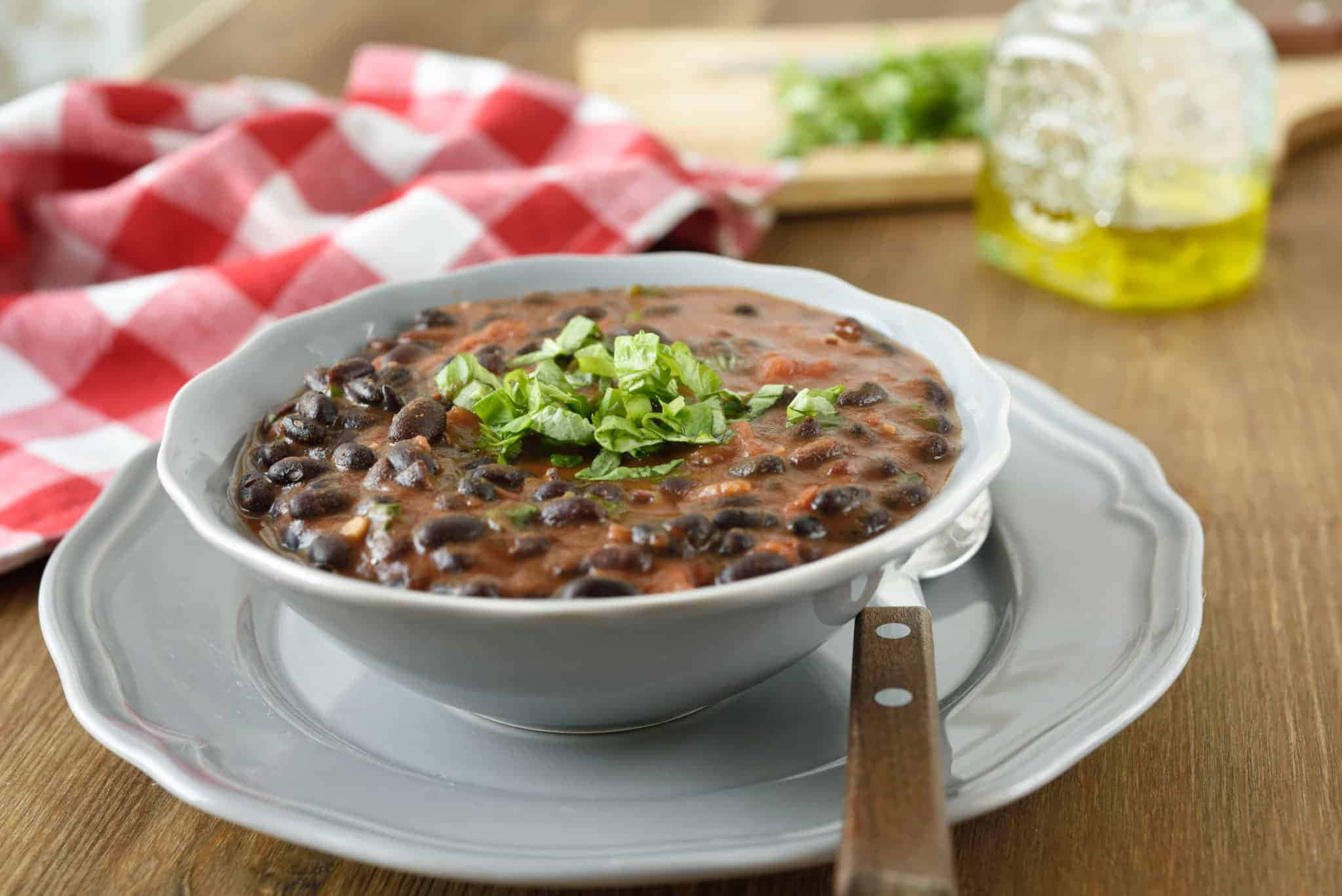 Black Bean Soup 桌布 壁紙 — 一碗冒煙的黑豆，上面放著新鮮芫荽，放在灰色碟子上，旁邊有一張紅白格仔餐巾。