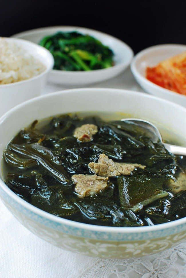 Seaweed 桌布 壁紙 — 一碗冒著熱氣的深色海帶湯，裡面有嫩肉塊，放在中央，周圍有米飯、泡菜和青菜等配菜。
