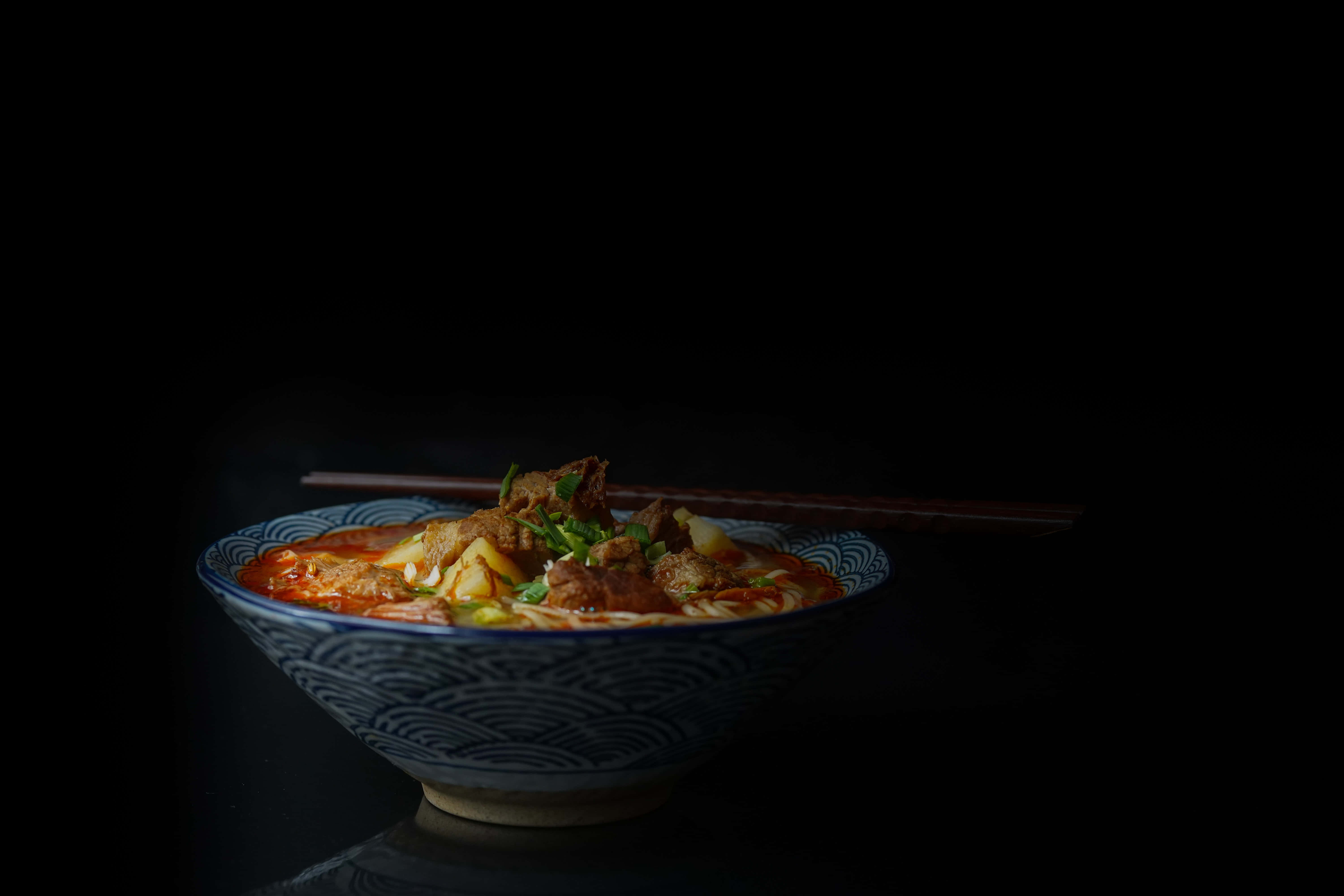 美食 桌布 壁紙 — 一碗冒著熱氣的濃郁紅色湯麵，配上嫩肉和青蔥，在戲劇性的聚光燈下，在漆黑的背景中閃耀。