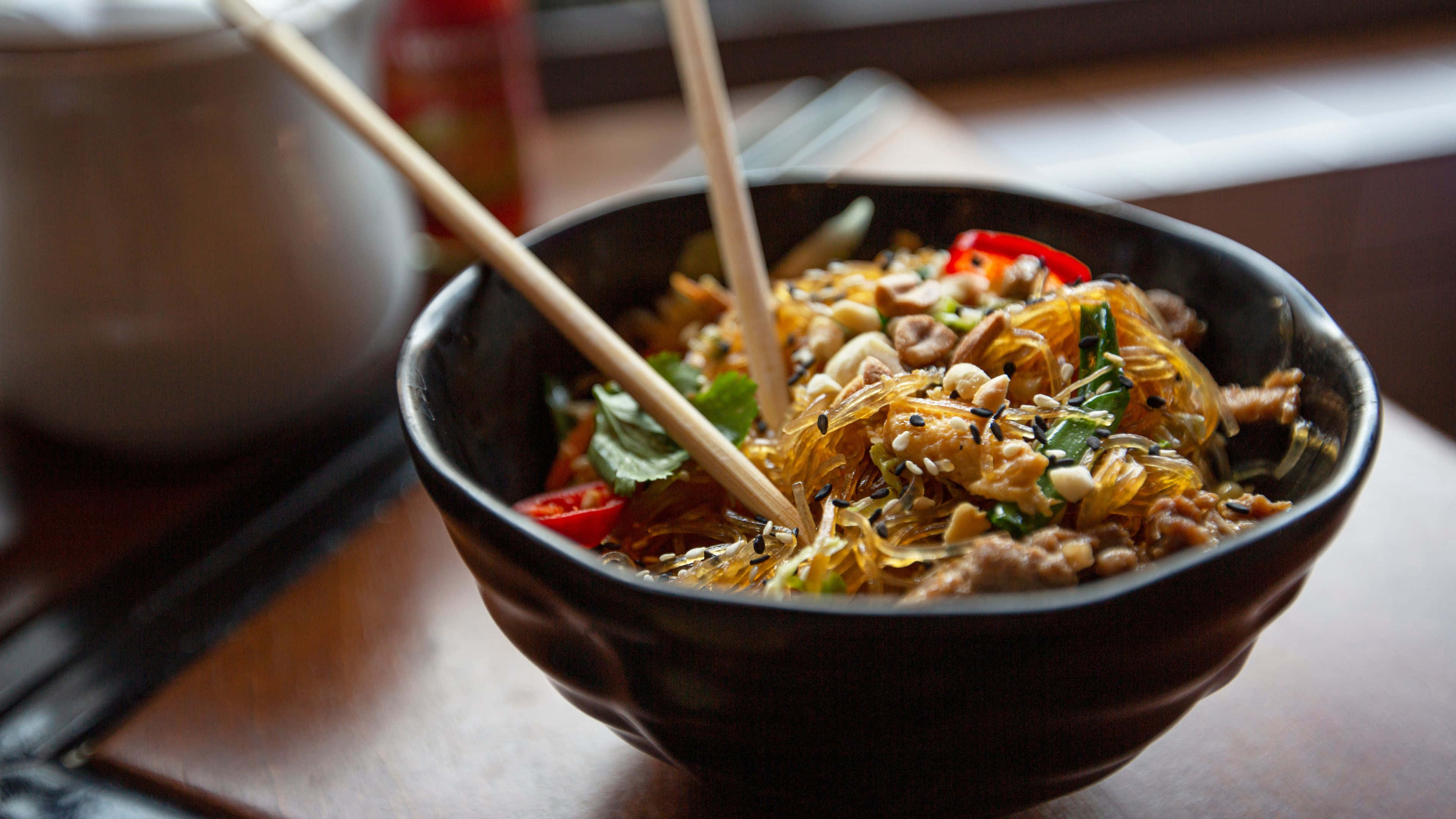 Noodles 桌布 壁紙 — 一碗冒著熱氣的辣味玻璃麵，配有肉、花生和香草，筷子擱在碗邊，隨時可以享用。