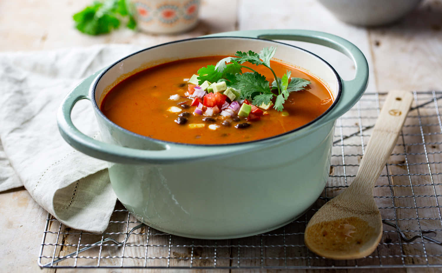Black Bean Soup 桌布 壁紙 — 一碗冒著熱氣的鮮橙色湯，上面放著新鮮芫茜、番茄粒和黑豆，放在金屬架上，旁邊有一把木湯匙。