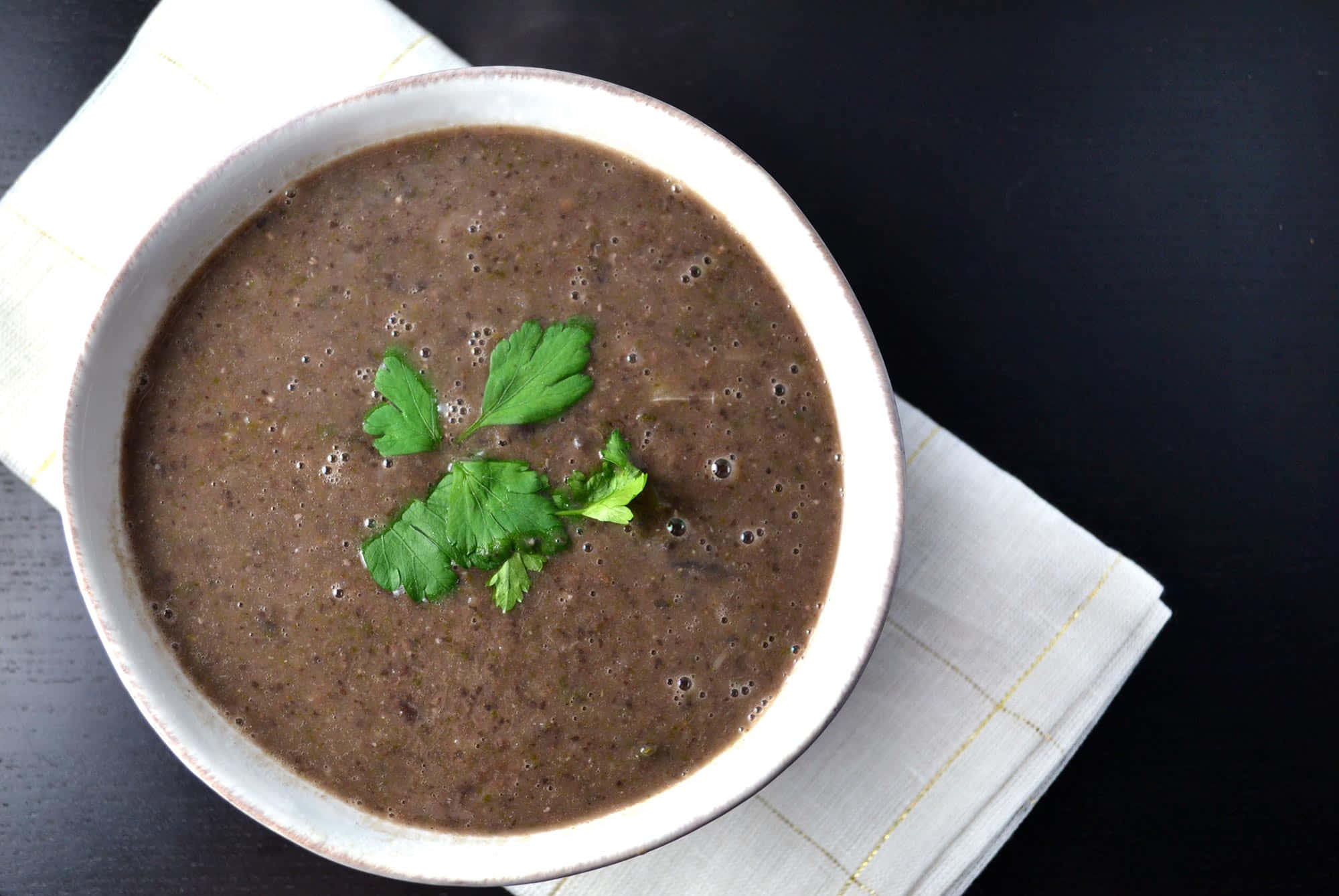 Black Bean Soup 桌布 壁紙 — 一碗奶油棕色湯放在白色碗中，飾以新鮮綠色歐芹，置於白色餐巾上，背景深色。