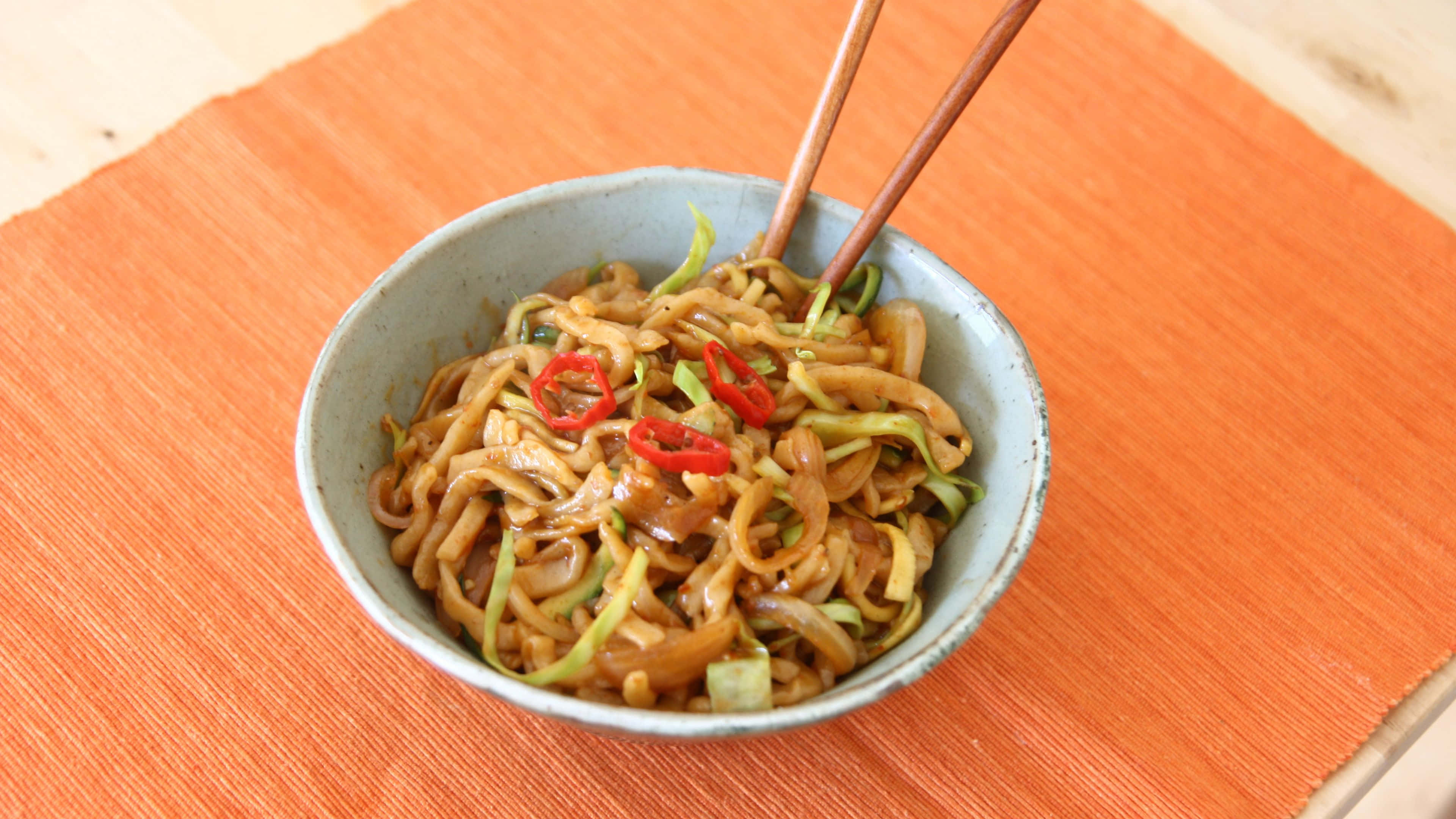 Noodles 桌布 壁紙 — 一碗油亮亮的炒麵，配上紅辣椒片和綠色蔬菜，放在橙色編織墊上，旁邊有木筷子。