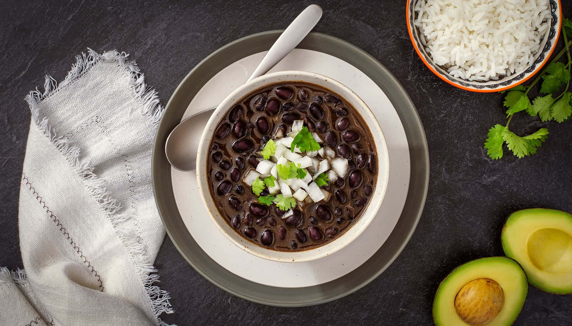 Black Bean Soup 桌布 壁紙 — 一碗濃郁的黑豆，上面放上新鮮芫荽和切碎的洋蔥，旁邊放著白飯、牛油果和亞麻餐巾，置於深色石板表面上。
