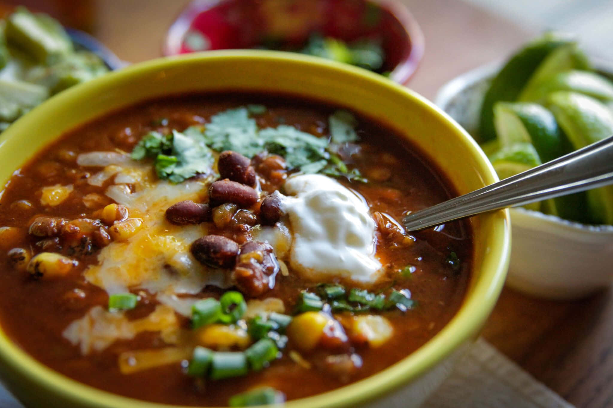 Black Bean Soup 桌布 壁紙 — 一碗熱氣騰騰的濃郁辣椒，上面放有酸奶油、香菜和青檸角，誘人地放在木桌上。
