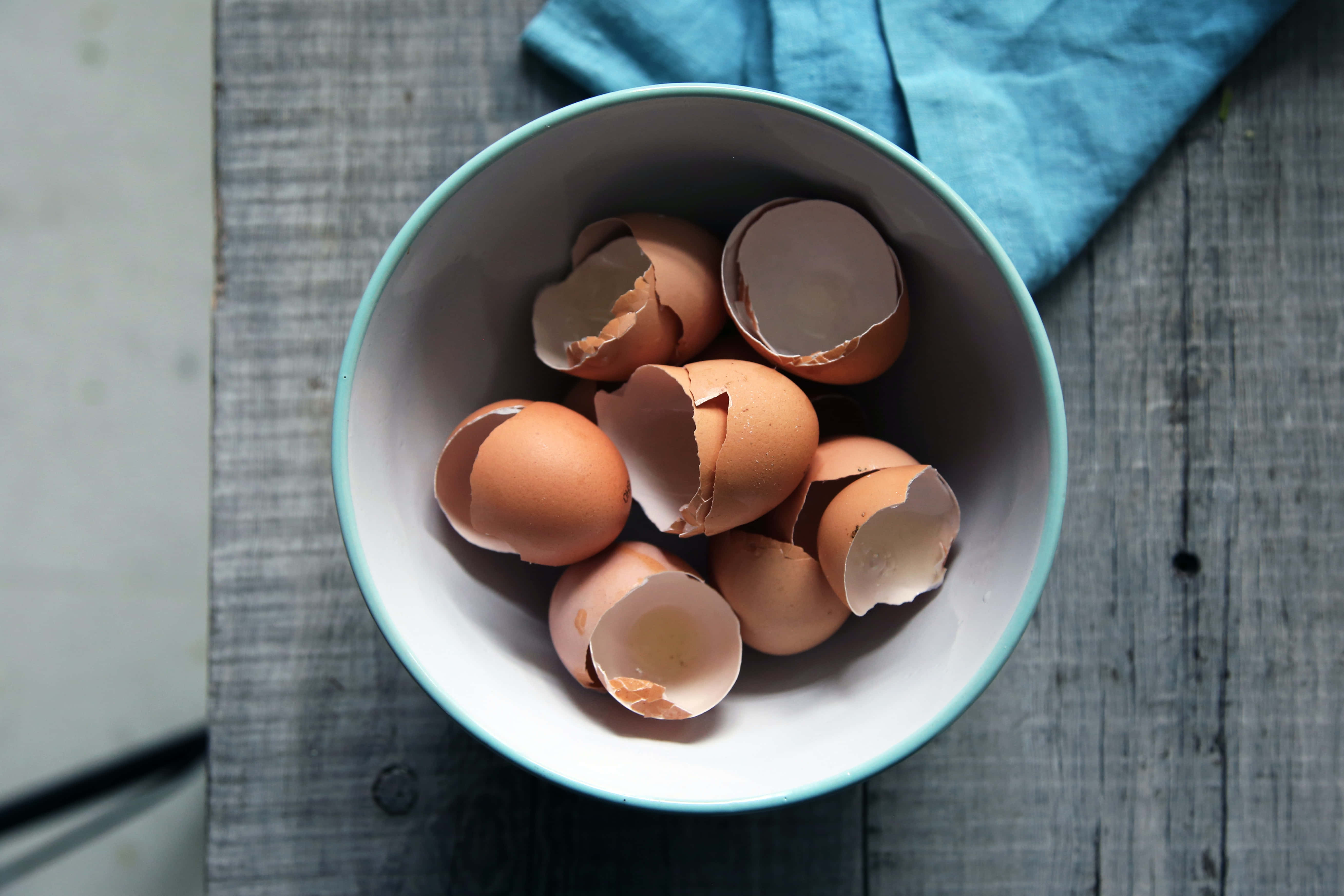 Brown Eggs 桌布 壁紙 — 一碗破碎的棕色蛋殼放在風化的木質表面上，旁邊有一塊藍布。