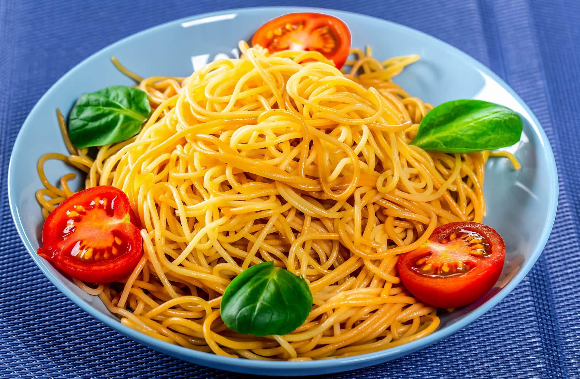 Noodles 桌布 壁紙 — 一碗色彩繽紛的意大利麵，上面放著新鮮羅勒和對半切開的櫻桃番茄，放在藍色紋理餐墊上。