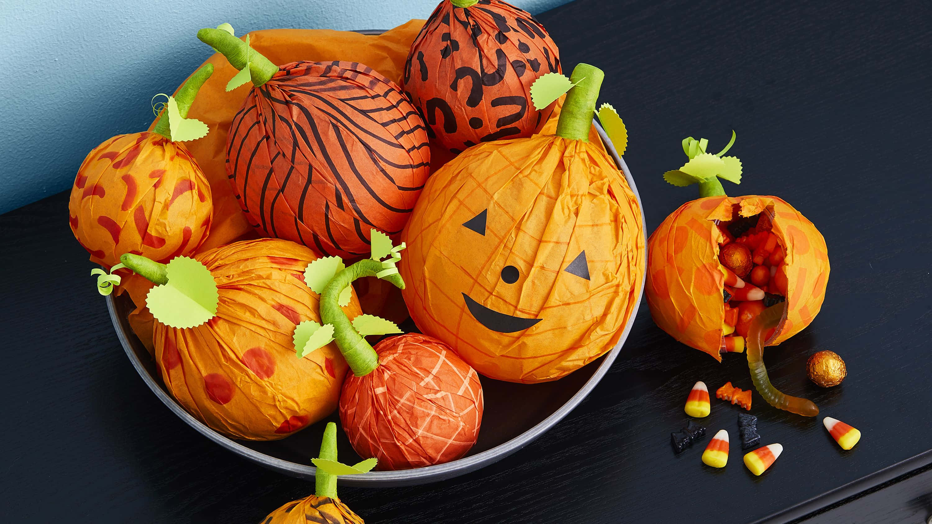 Halloween Props 桌布 壁紙 — 一碗色彩繽紛的紙製萬聖節南瓜，糖果灑落在深色桌子上。