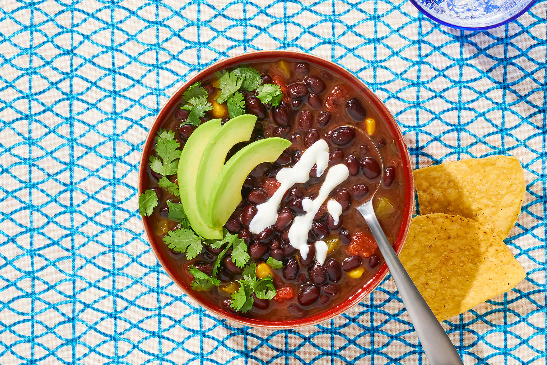 Black Bean Soup 桌布 壁紙 — 一碗色彩繽紛的黑豆辣椒，上面放著牛油果片、酸忌廉和芫荽，放在藍色圖案枱布上，附近有墨西哥玉米片。