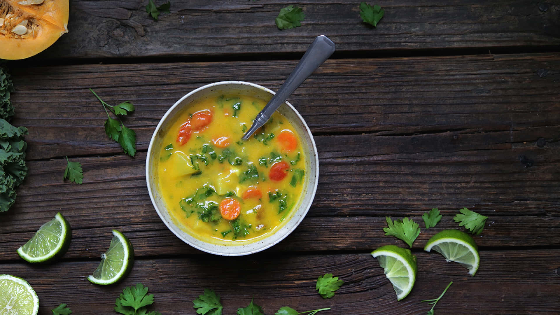Soup 桌布 壁紙 — 一碗色彩鮮豔的蔬菜湯，有紅蘿蔔和芫荽，放在深色木桌上，周圍有青檸角和新鮮香草。