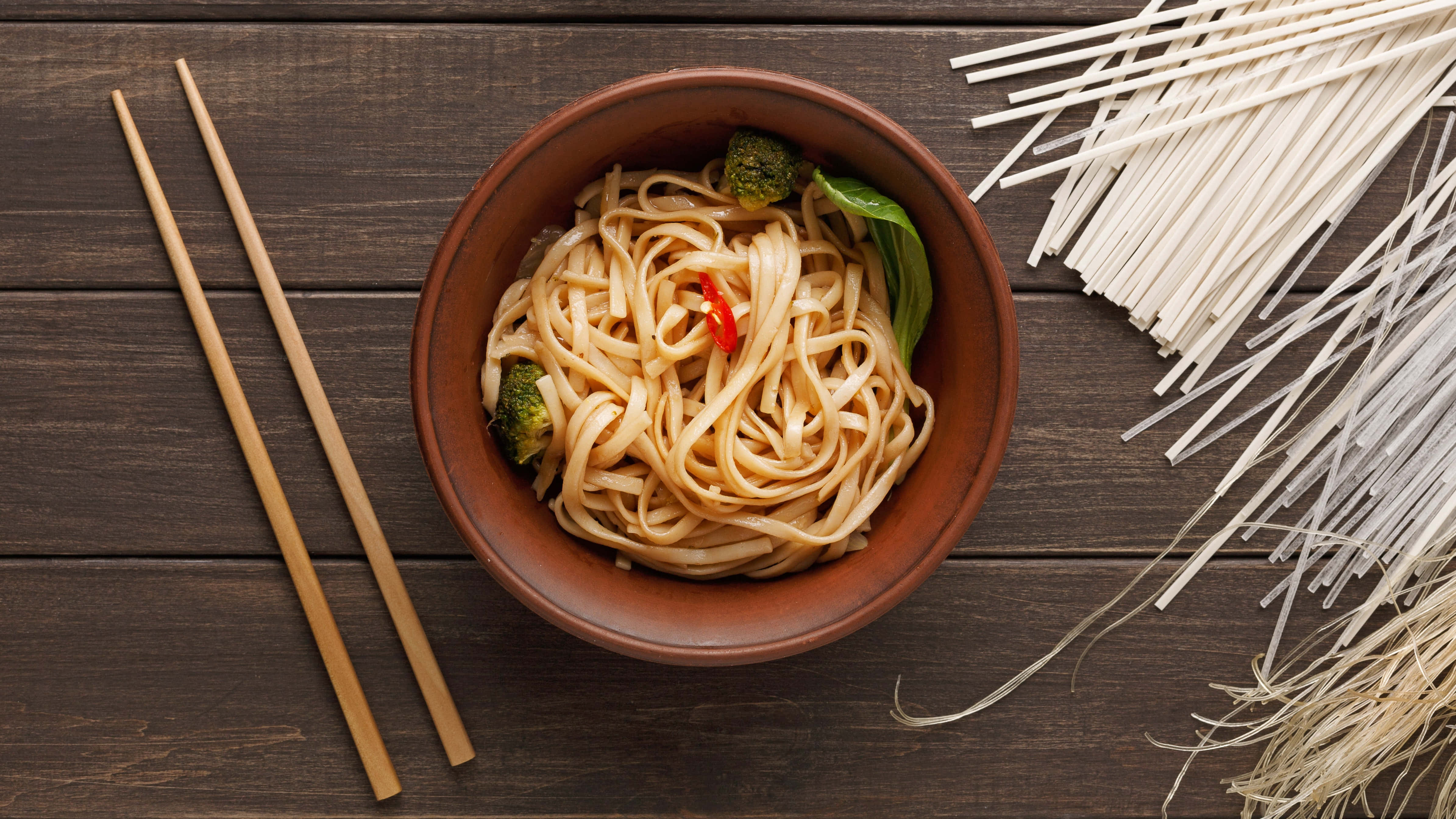 Noodles 桌布 壁紙 — 一碗鄉村風格的麵條配西蘭花和辣椒，旁邊放著生麵條和筷子，放在深色木桌上。