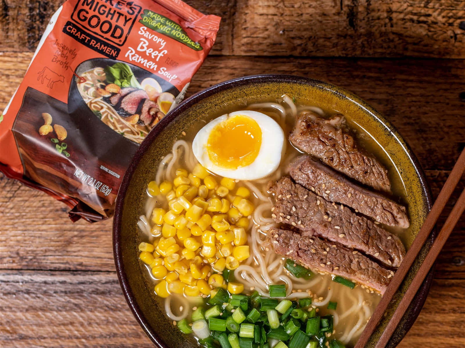 一碗香濃牛肉拉麵湯 壁紙 — 一碗冒著熱氣、滋味濃郁的牛肉拉麵，上面放著半熟蛋、粟米和青蔥，旁邊放著調味包，擱在質樸的木桌上。