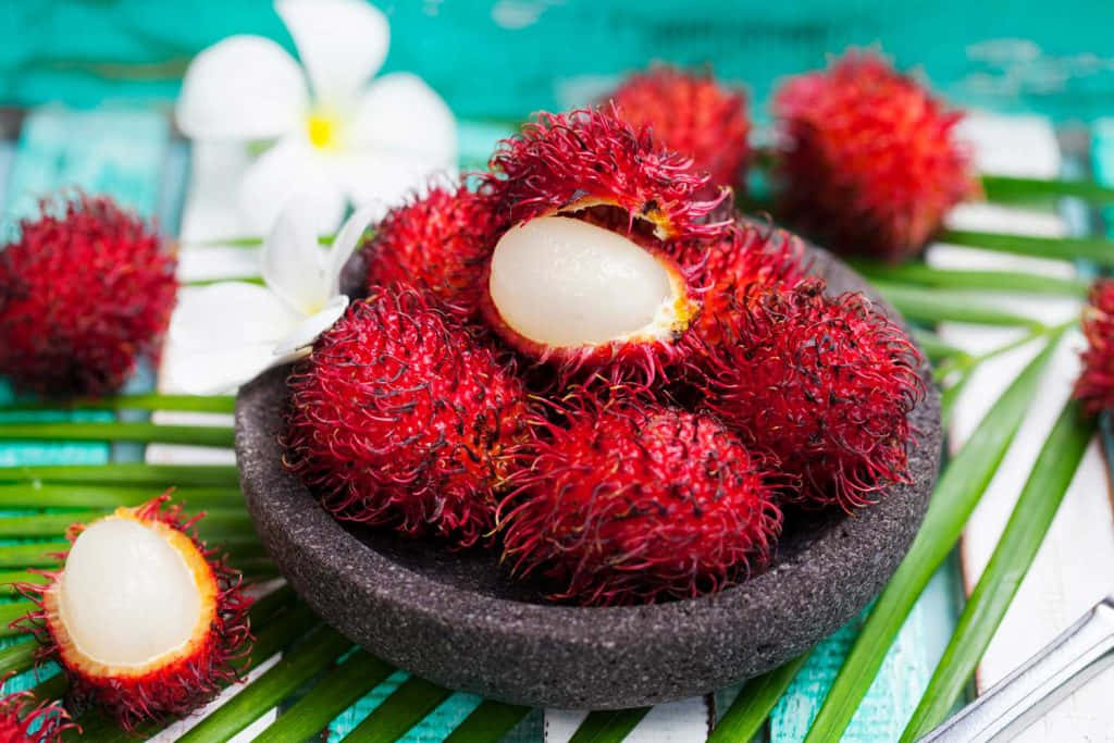 Red Fruit 桌布 壁紙 — 一碗鮮紅色、果肉白色的紅毛丹放在綠葉上，背景是藍綠色的木質表面。
