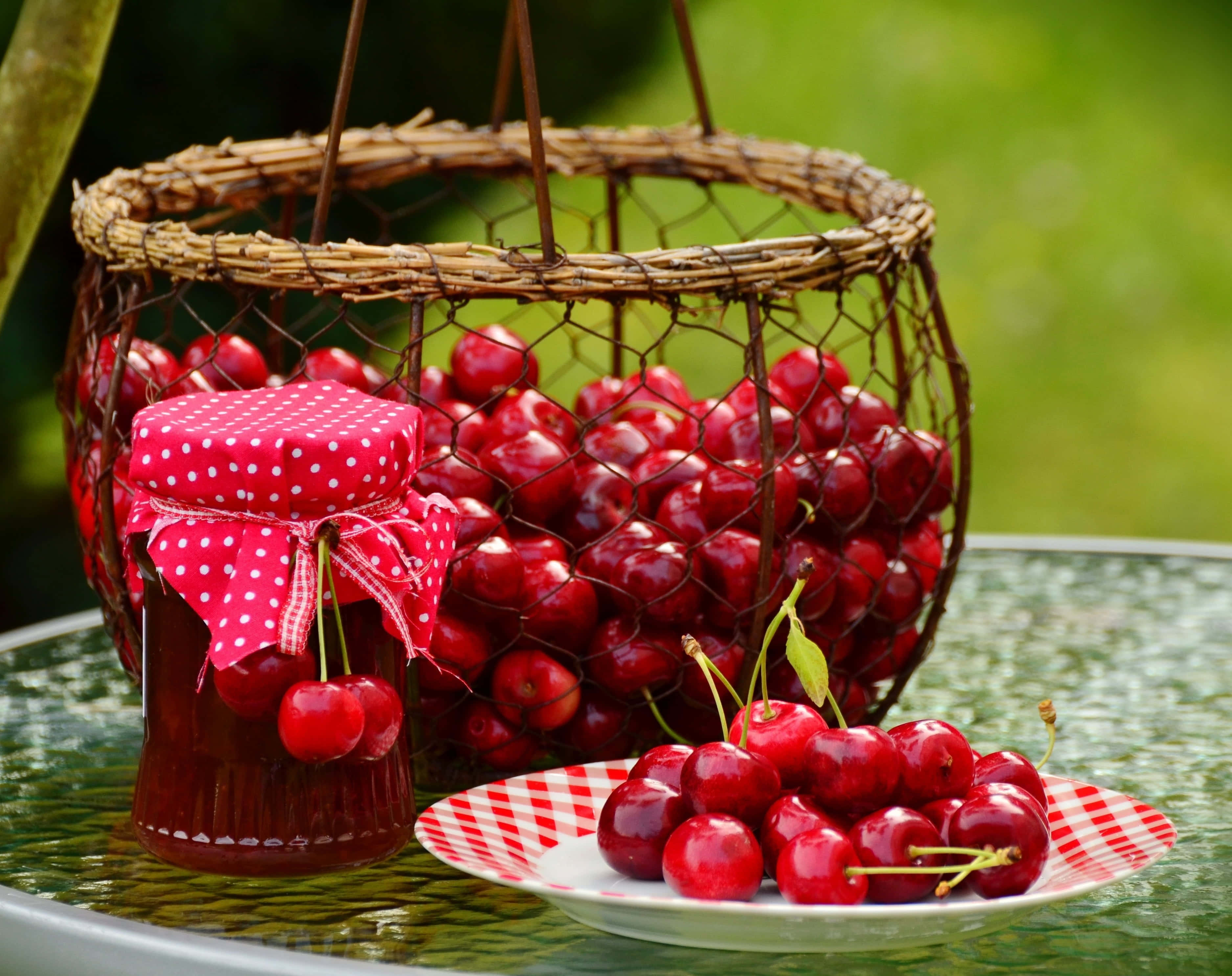 Spring Desserts 桌布 壁紙 — 一籃子光澤的紅櫻桃旁邊放著一瓶櫻桃果醬和一盤新鮮櫻桃，放在戶外的玻璃桌上。