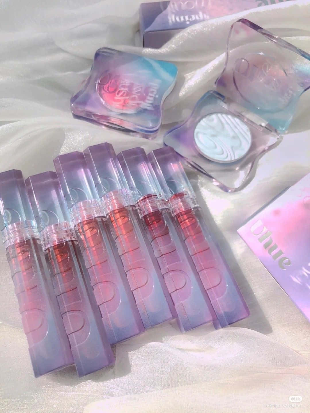 lipstick 桌布 壁紙 — 一系列彩虹色調的紫色和粉紅色唇彩和粉末，排列在柔軟的白色布料上，帶有夢幻般的柔和光澤。