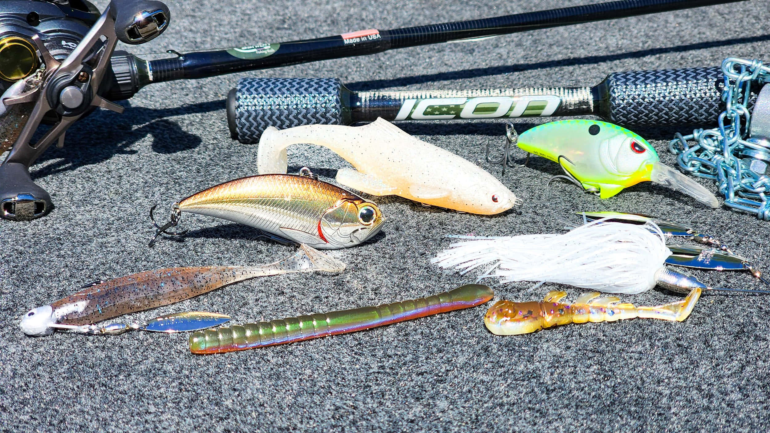 Fishing Bait 桌布 壁紙 — 一系列色彩繽紛的魚餌和一支魚竿放在有紋理的灰色瀝青上，在明亮的陽光下。