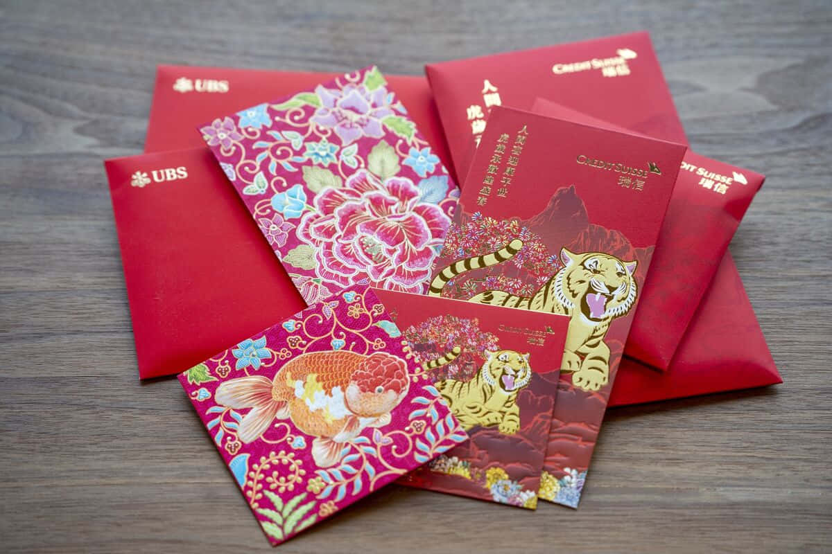 Red Envelope 桌布 壁紙 — 一系列色彩鮮豔的紅色和粉紅色中國新年利是封，帶有金色老虎、魚和花卉圖案，放置在木質表面上。