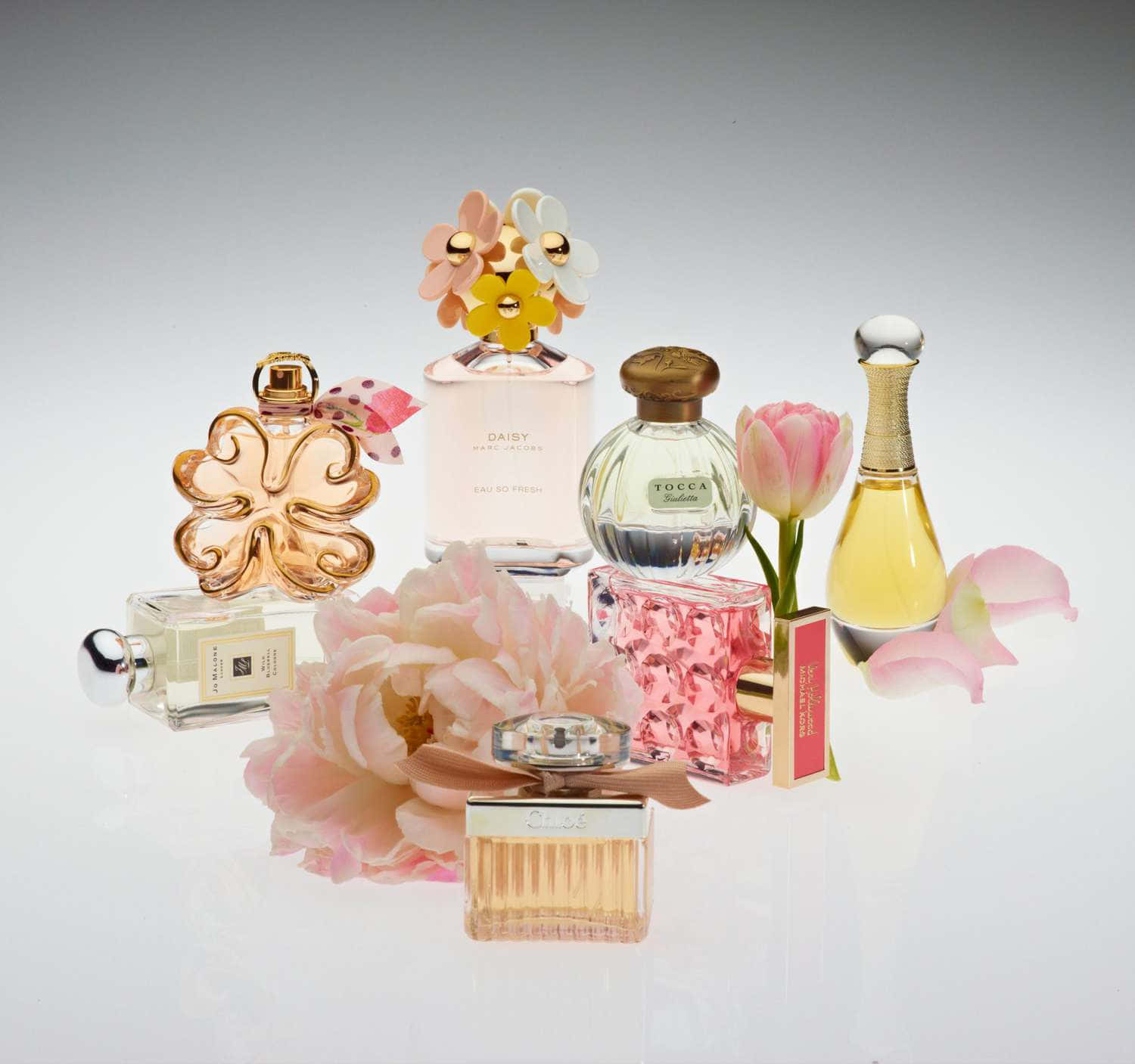 Floral Fragrance 桌布 壁紙 — 一系列迷人的設計師香水瓶，搭配粉紅色花朵，擺放在反光的白色表面上。