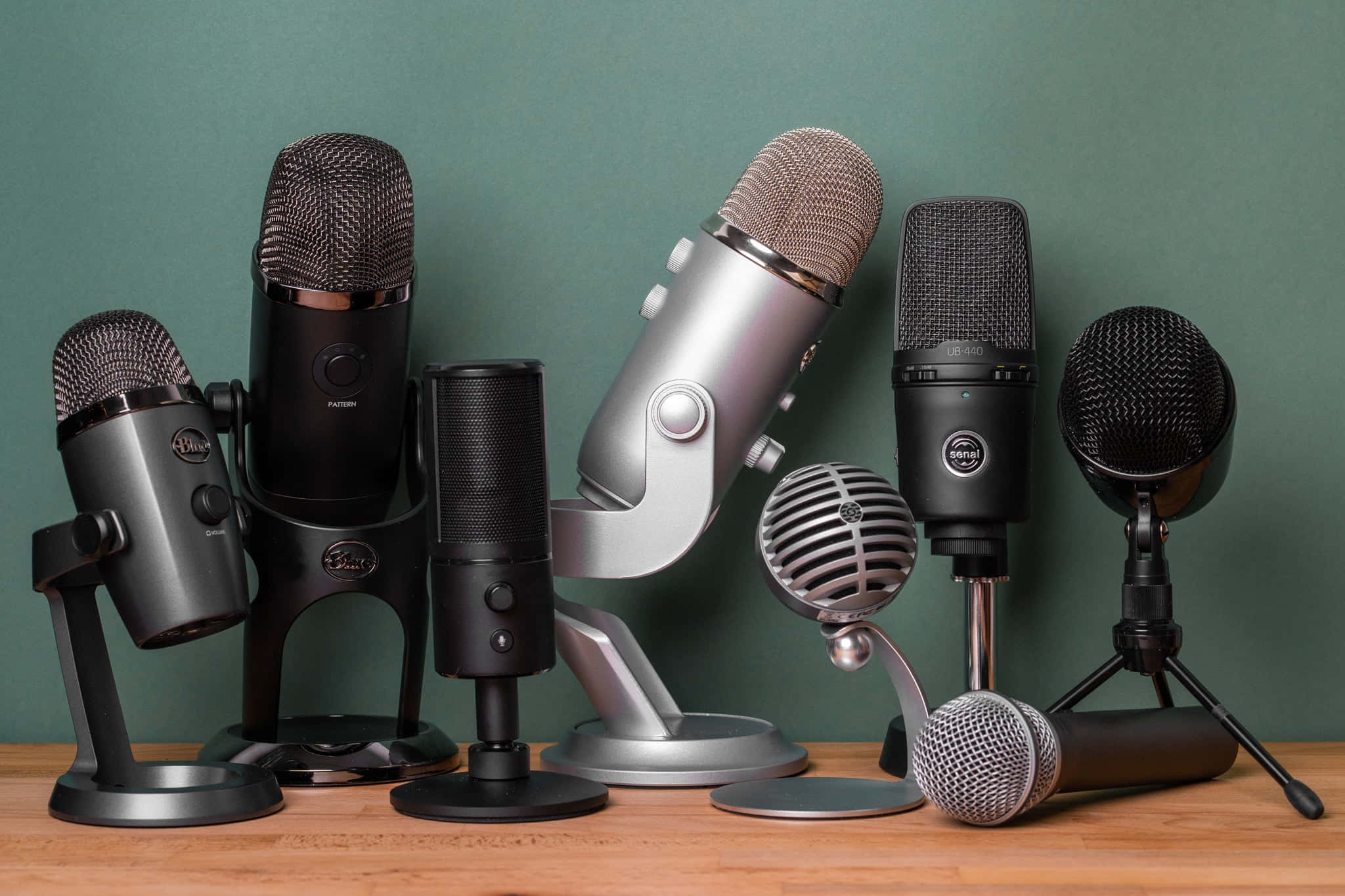 Microphone 桌布 壁紙 — 一系列黑色和銀色的專業麥克風排列在木質表面上，背景是柔和的綠色牆壁。
