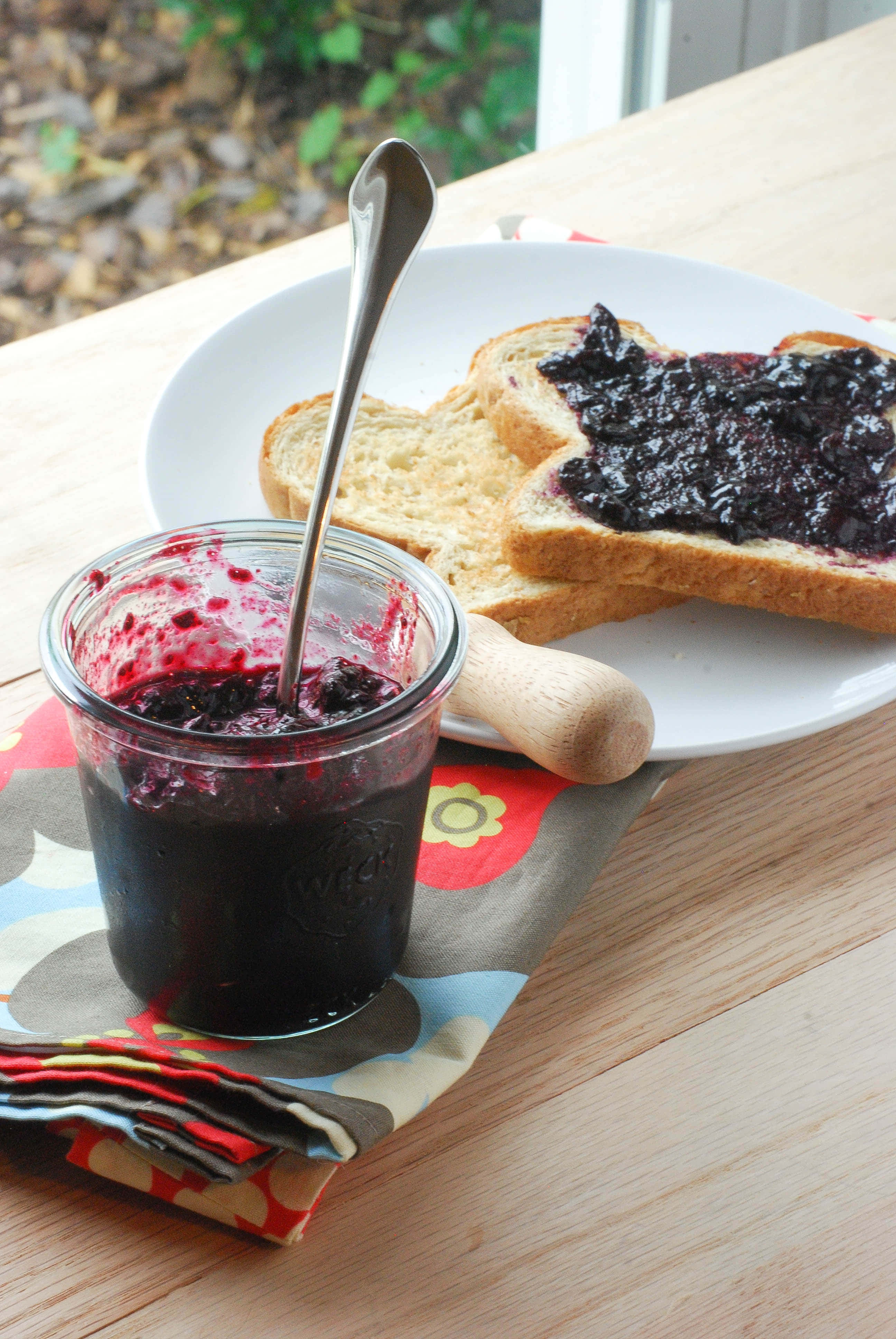 Blueberry Jam 桌布 壁紙 — 一罐深色莓果醬配湯匙，放在木桌上色彩繽紛的餐巾上的碟子旁，旁邊有多士。